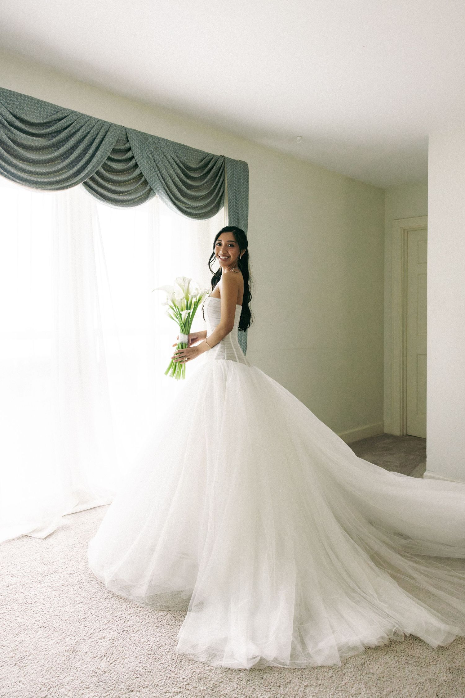 Bride in a flowing white ballgown holding a tulip bouquet stands by a draped window in a softly lit room.
