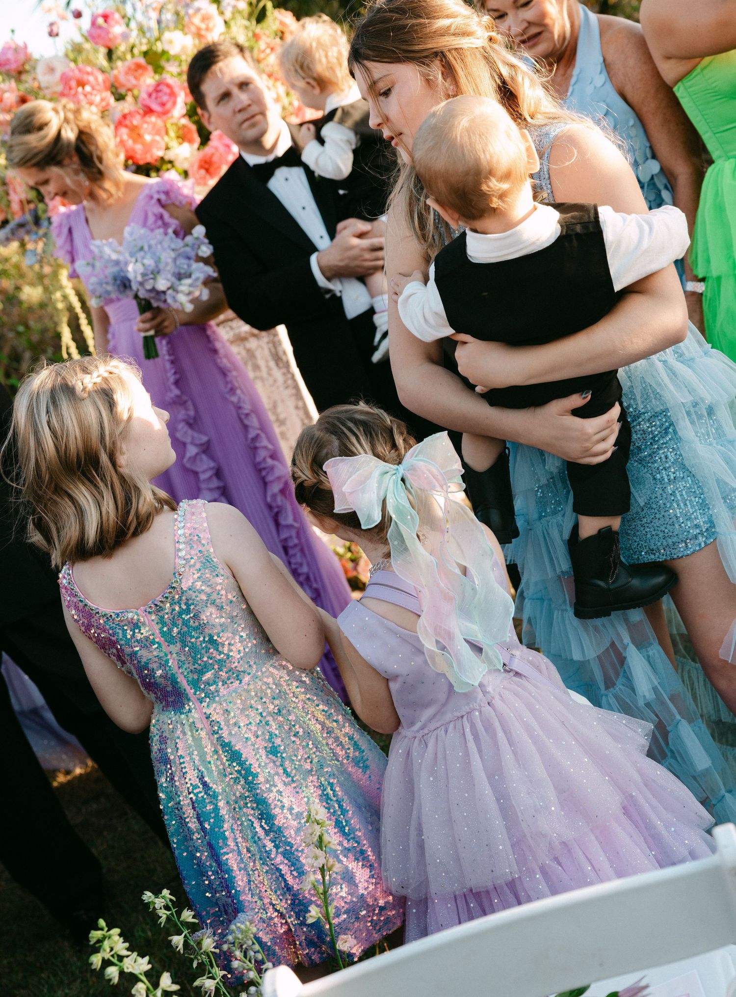 Children in colorful dresses mingle with wedding guests at an outdoor garden ceremony.
