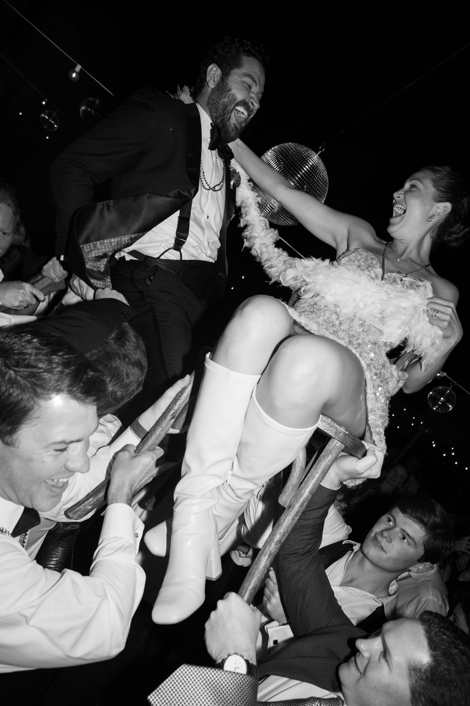 Bride and groom laughing while being lifted on chairs during a lively wedding hora dance celebration.