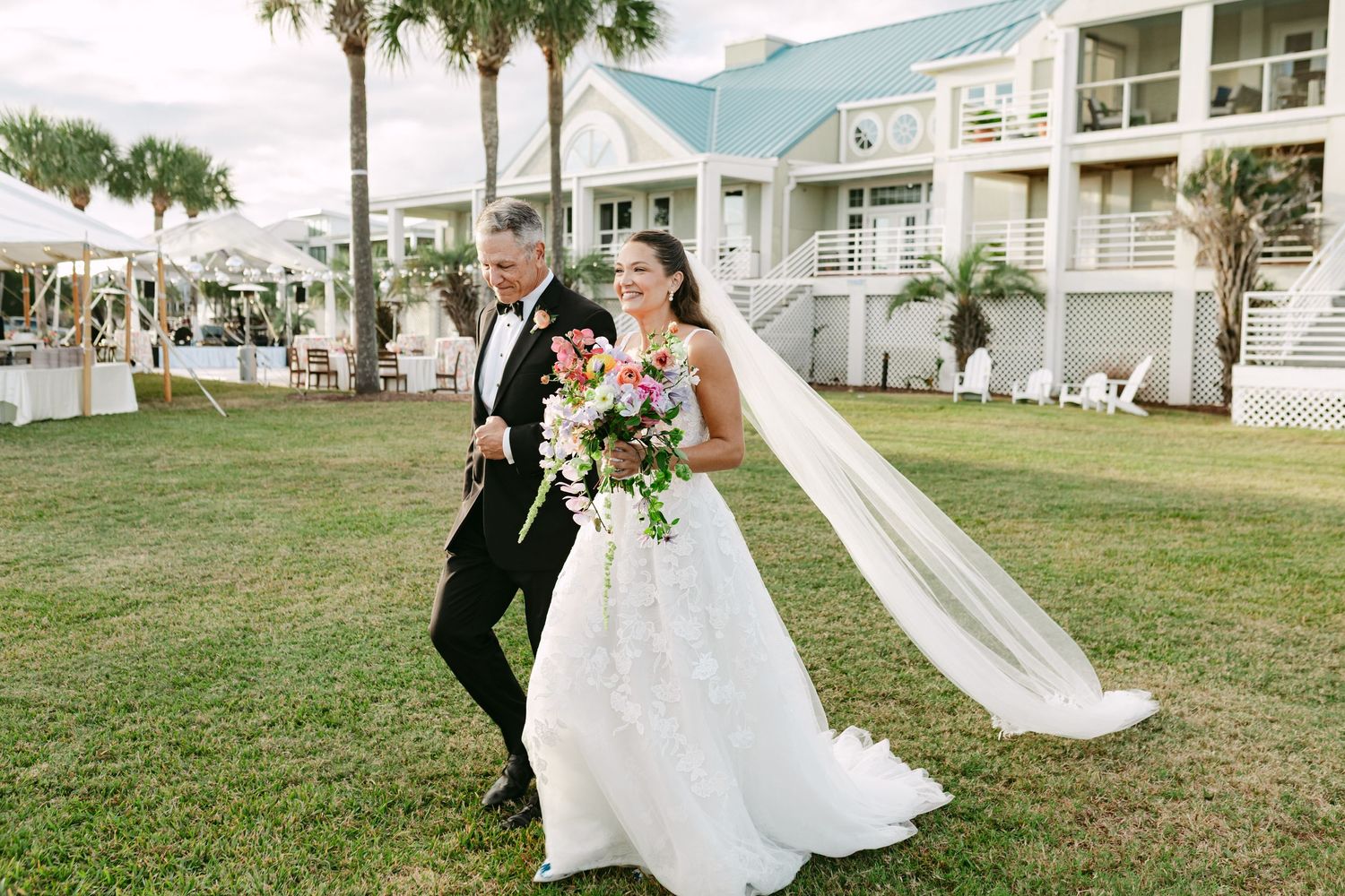 Bride in lace gown and long veil walks with escort near a coastal wedding venue.