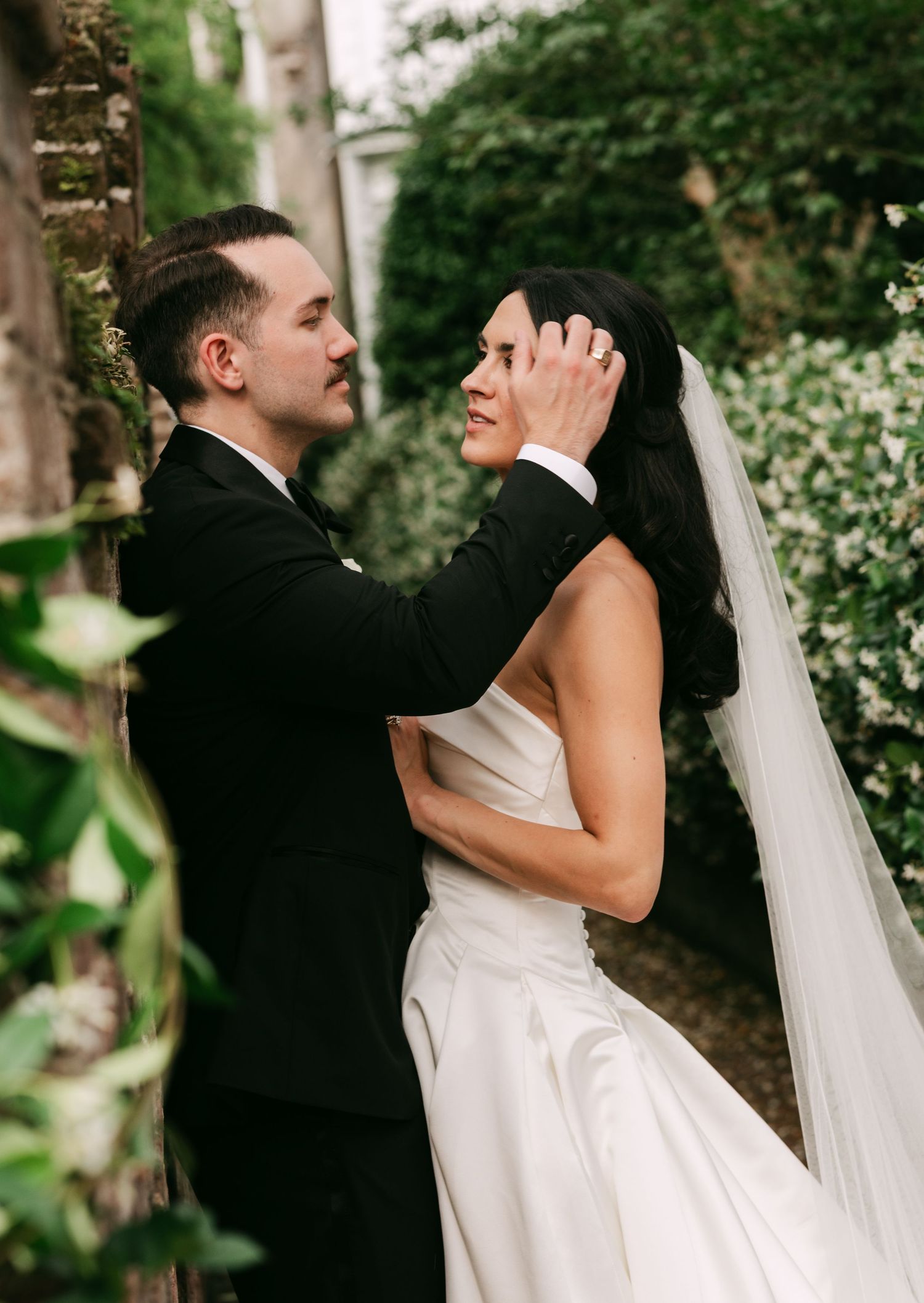 Groom in black tuxedo tenderly touches bride's face as she smiles in a white gown and veil outdoors.