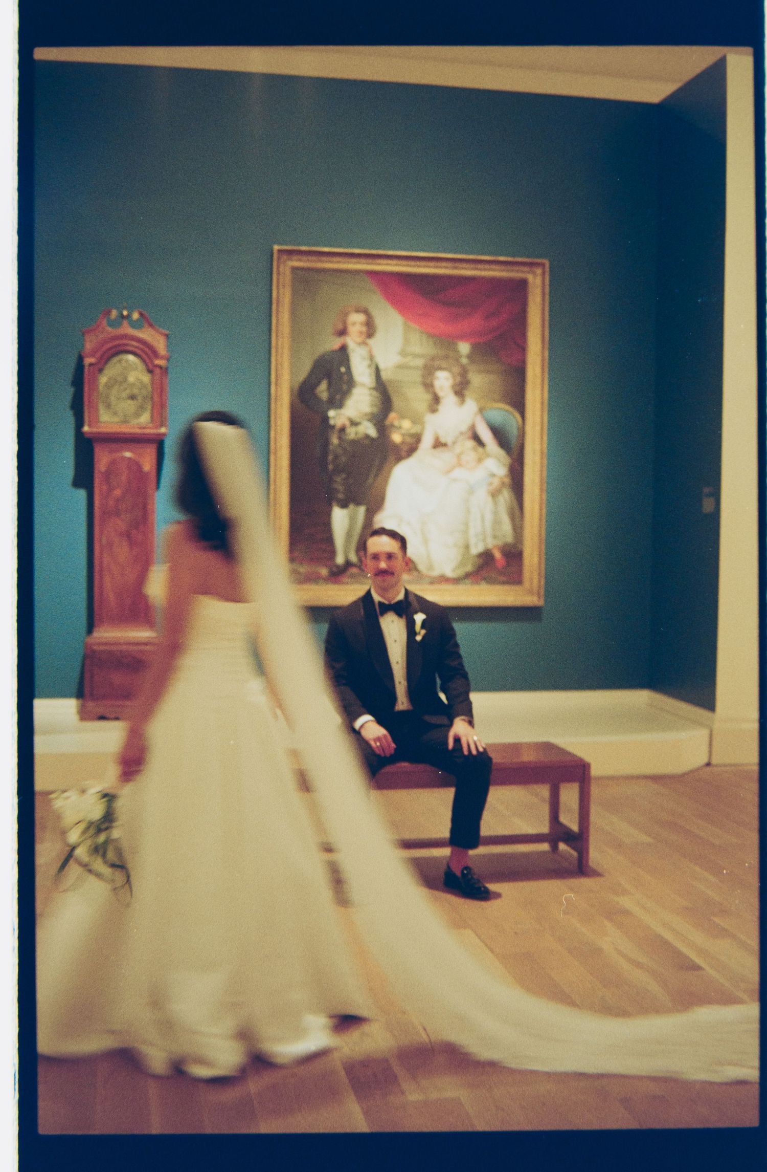 Groom seated on bench smiling as bride walks past in art museum gallery with colonial family portrait behind him.