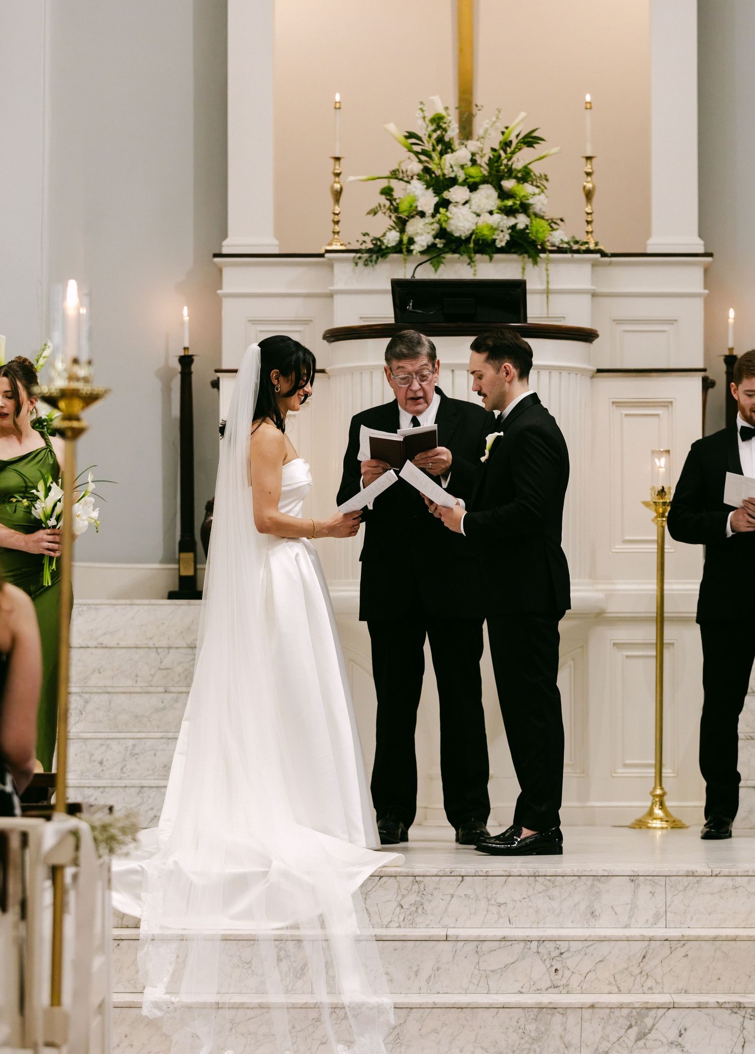 Bride and groom exchange vows at elegant church altar with white floral arrangements and candles.