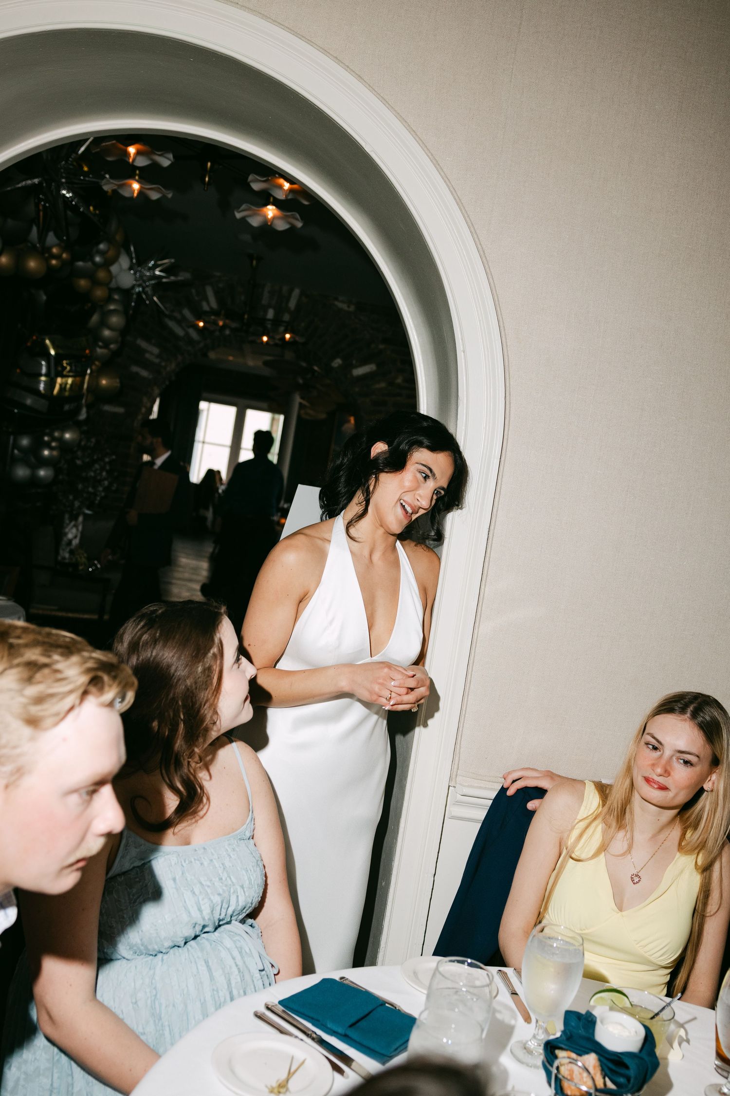 Bride in white halter gown mingles and laughs with seated wedding guests at the reception.
