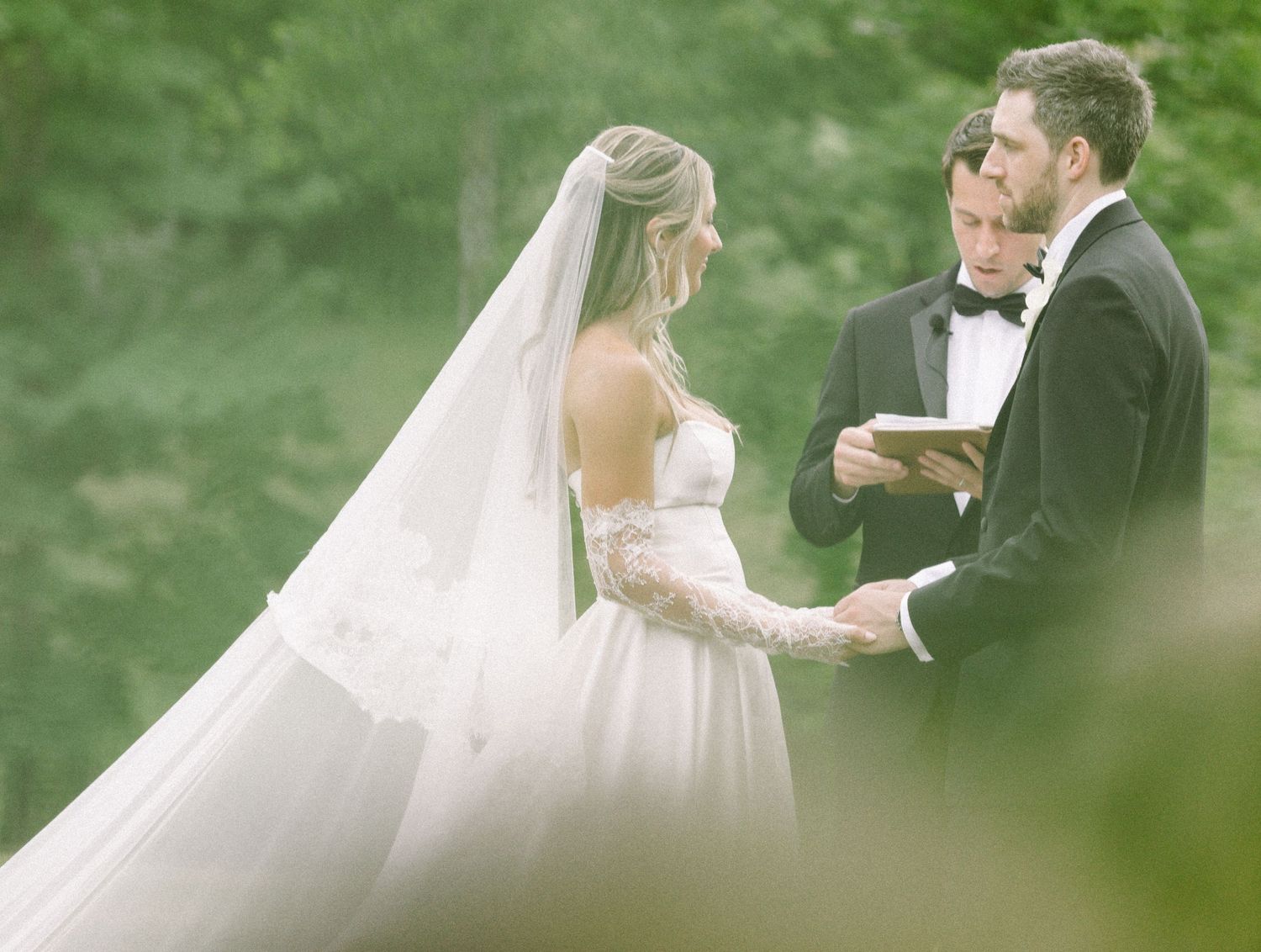 Bride in white gown and veil exchanges vows with groom in black tuxedo during outdoor wedding ceremony.