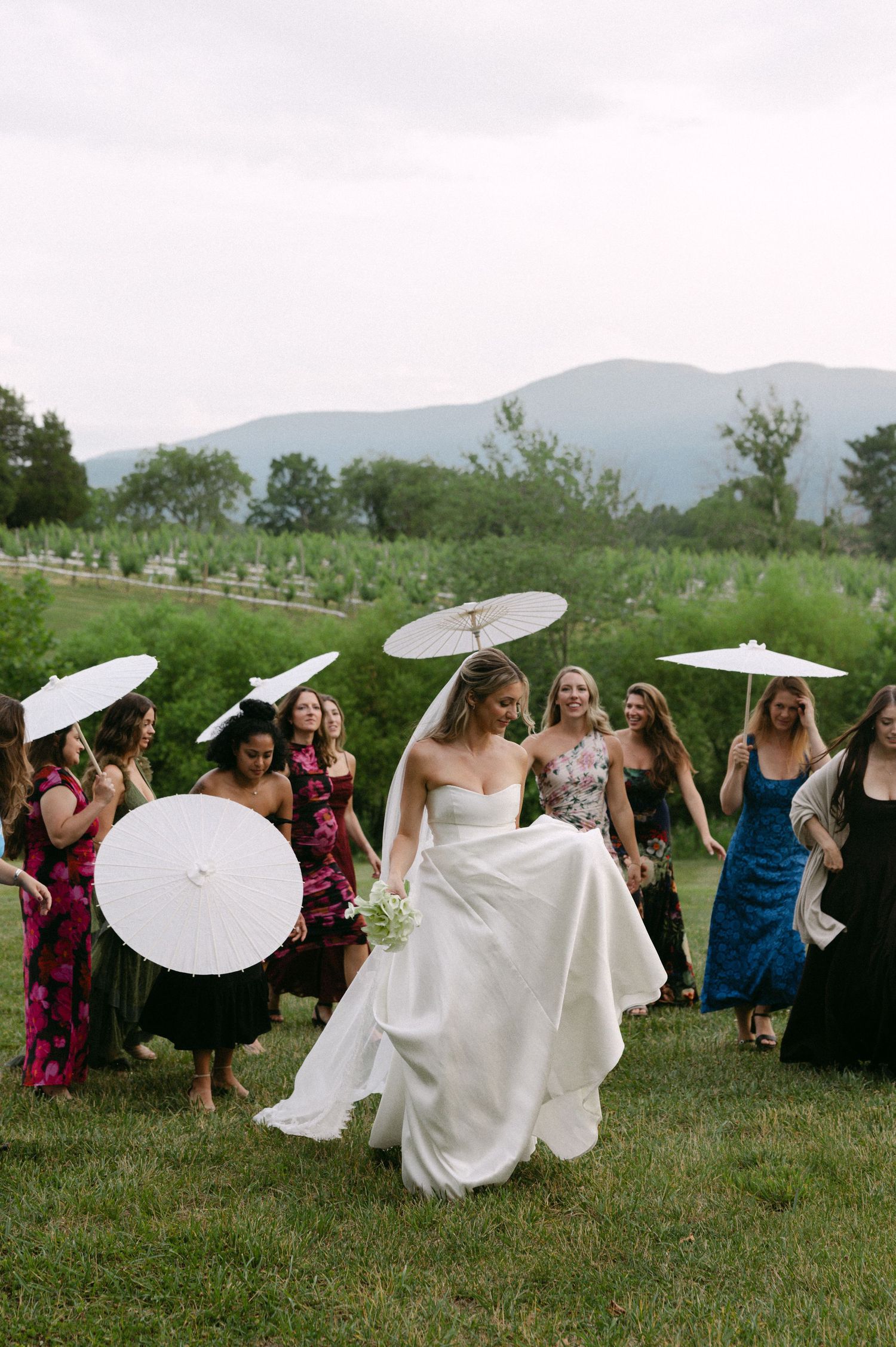 Bride in strapless white gown walks with bridesmaids holding white parasols on green lawn with mountain backdrop.