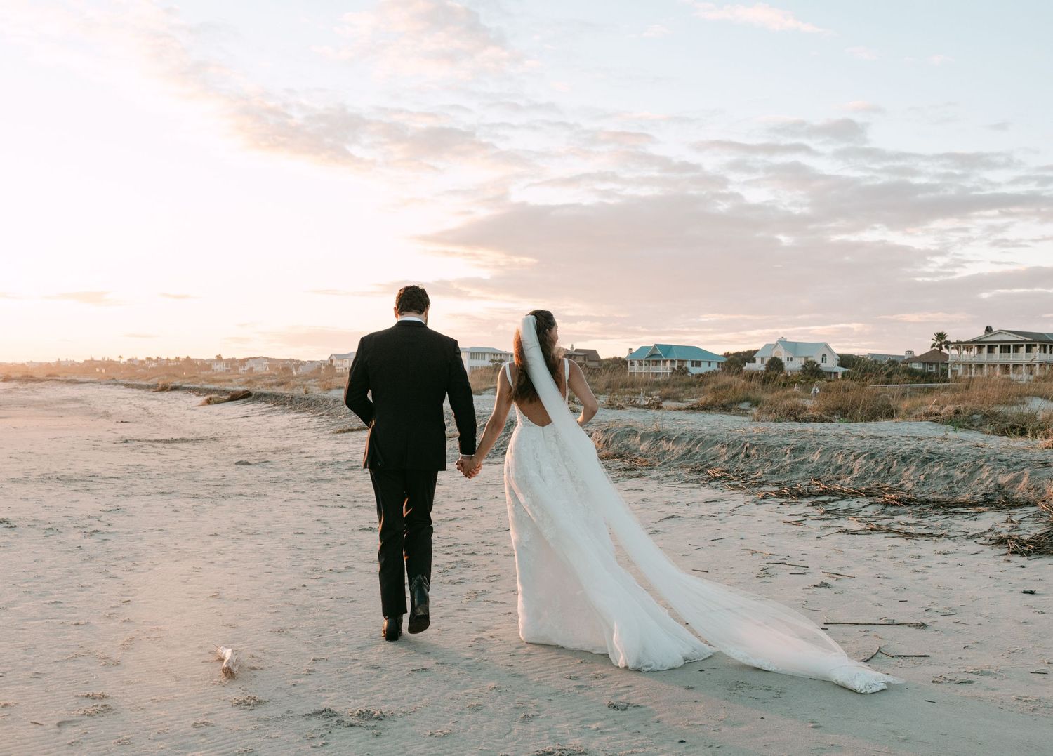 Bride and groom holding hands walking on beach at sunset with coastal homes in background.
