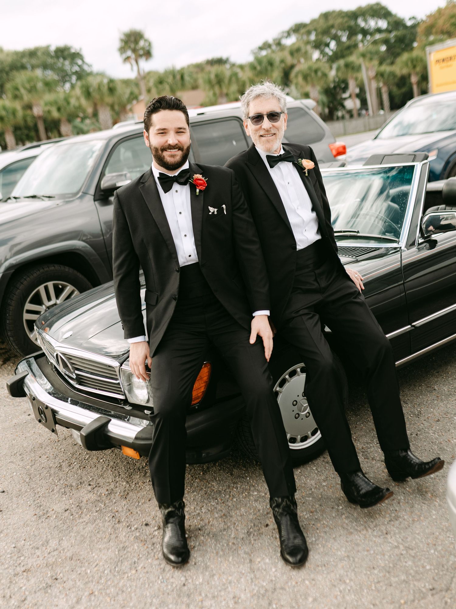 Groom and older man in matching black tuxedos pose smiling beside a vintage classic car outdoors.