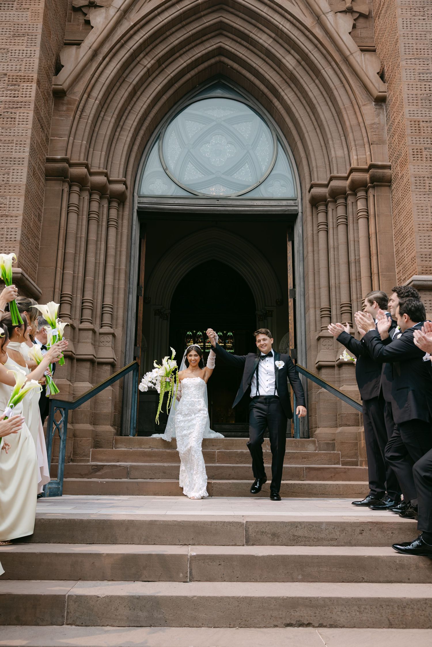 Joyful wedding party celebrates on church steps with raised bouquets.