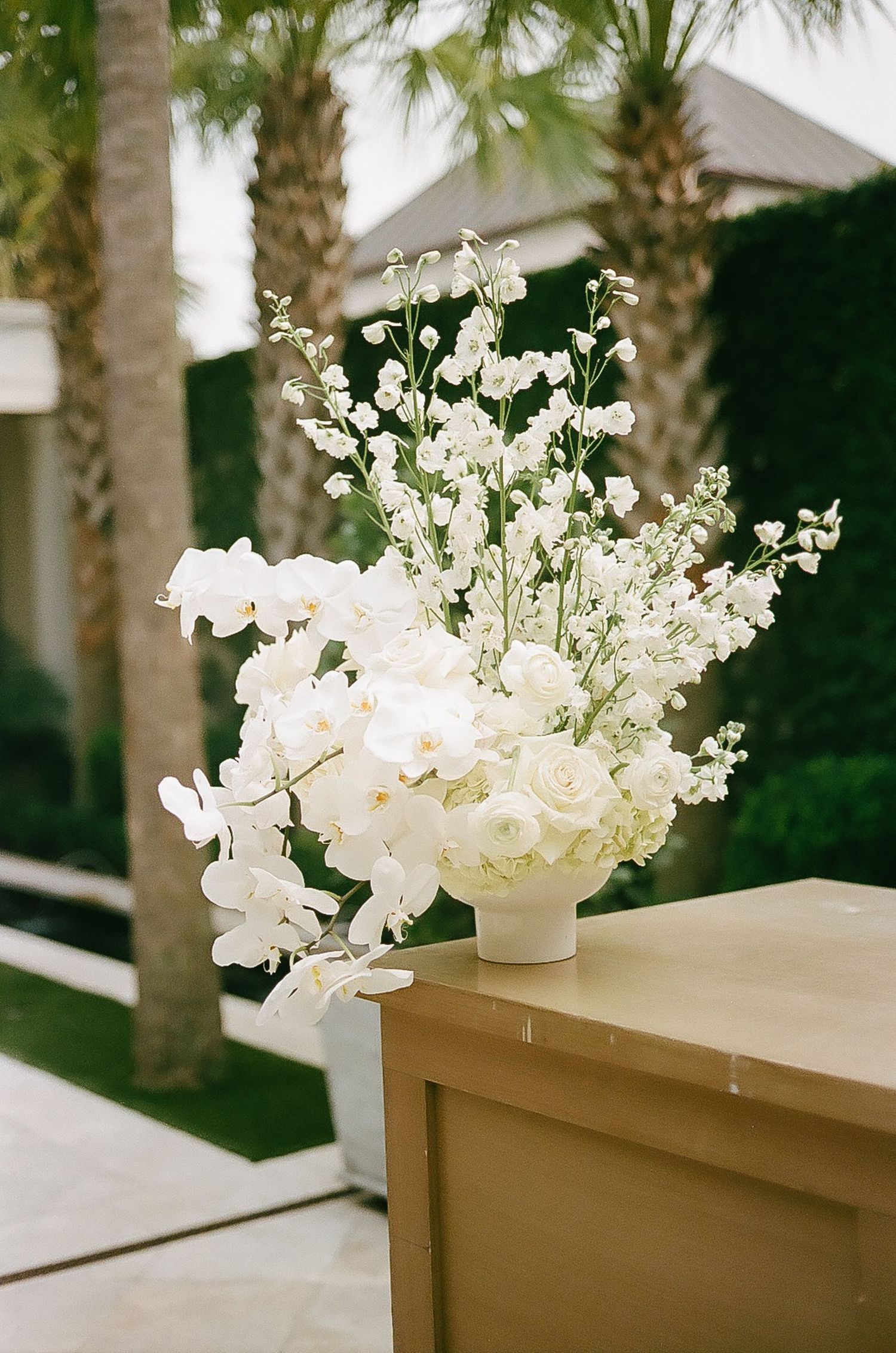 White floral centerpieces with orchids line a wooden table under palm trees at an outdoor venue.
