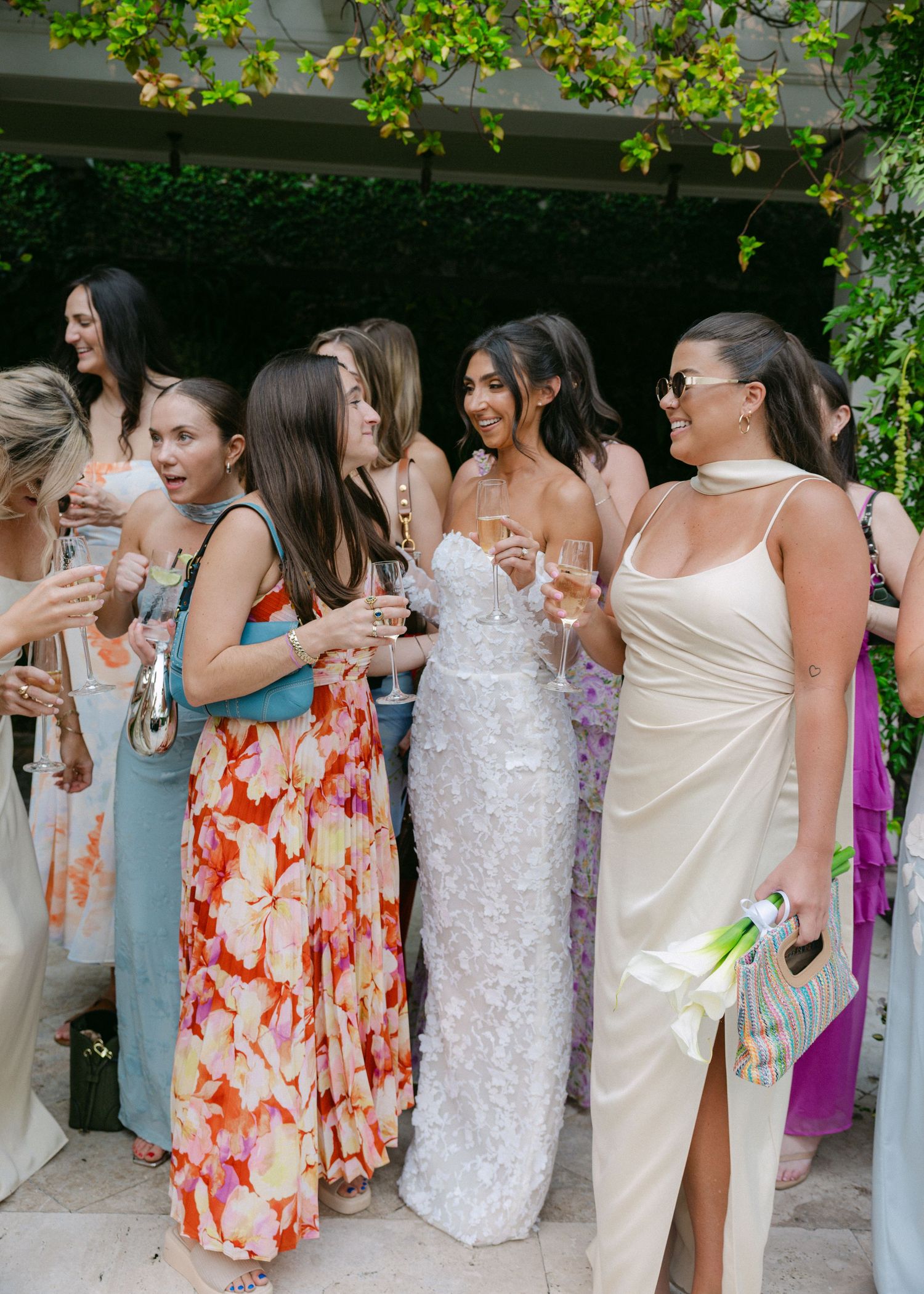Wedding guests in pastel and floral dresses gather under a vine-covered pergola.