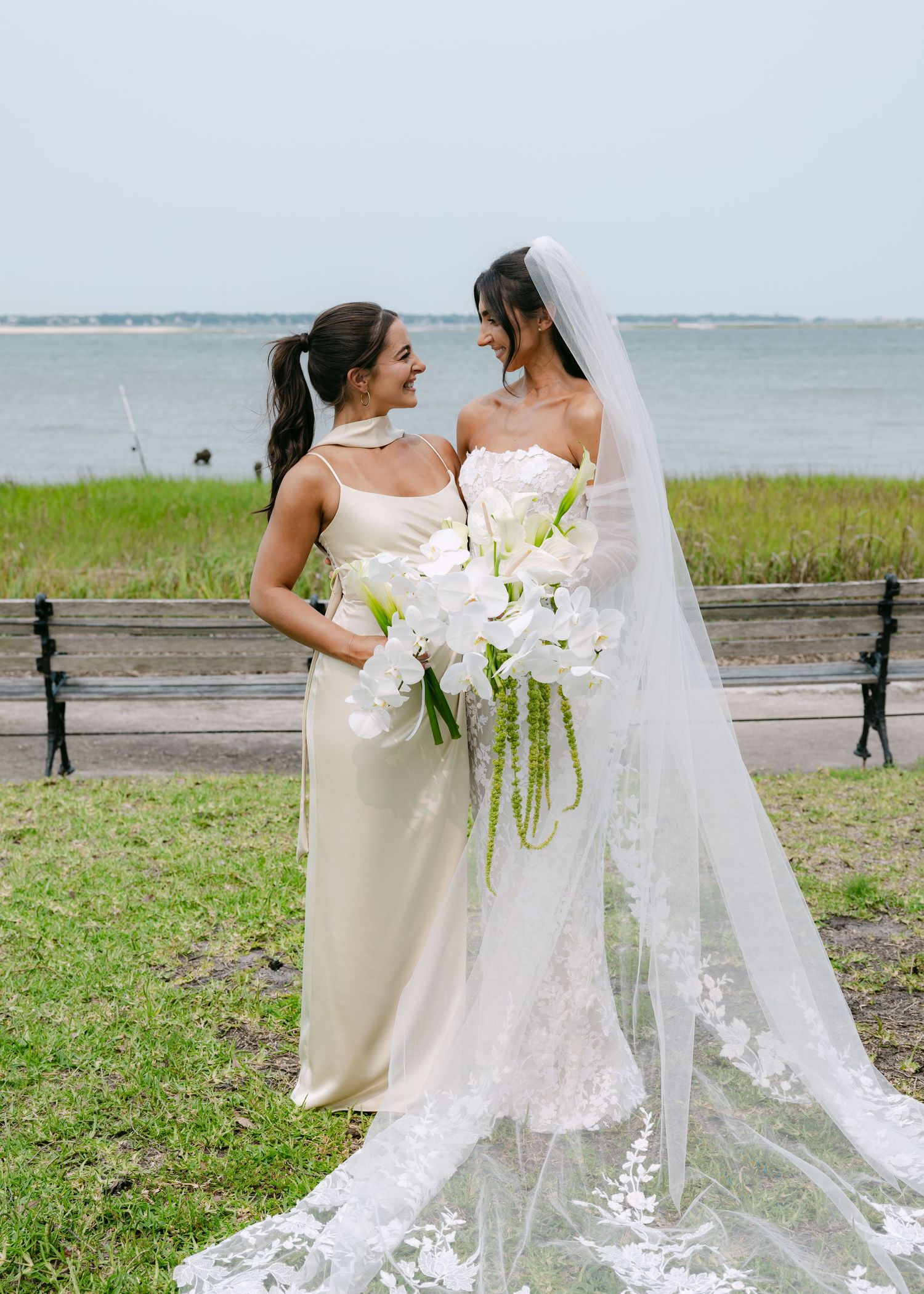 Two elegant wedding attendees embrace on a scenic grassy lawn by the ocean.