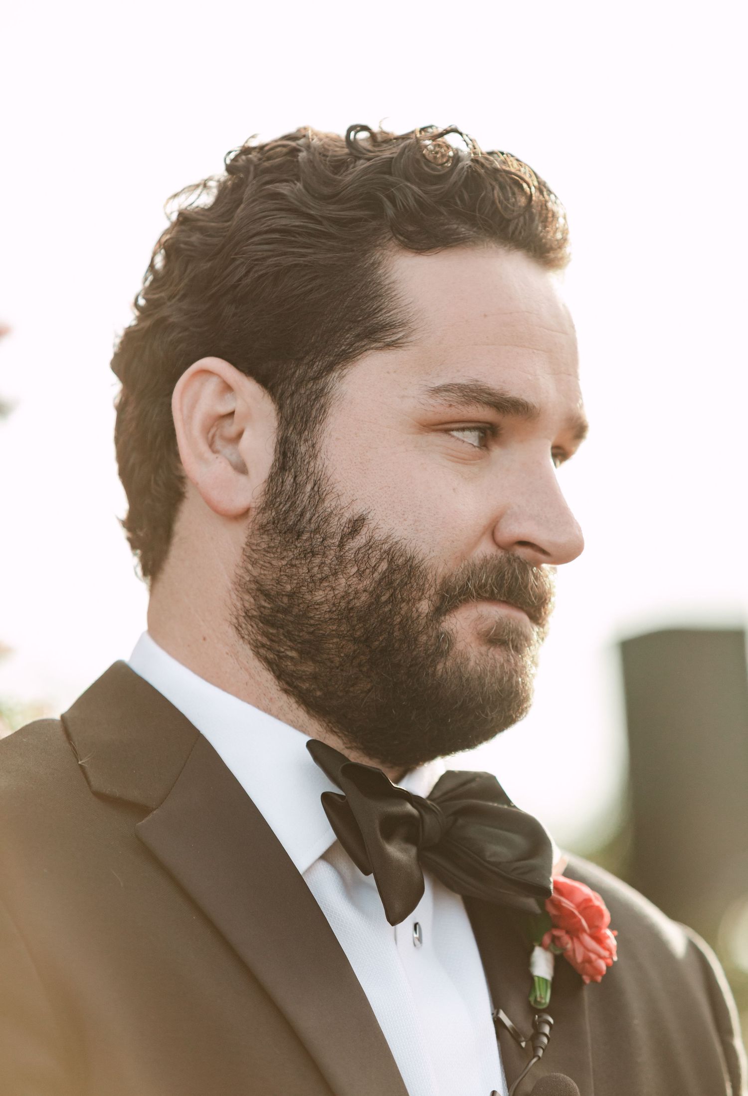 Bearded groom in black tuxedo and bow tie with a red boutonniere, photographed in profile at outdoor wedding.