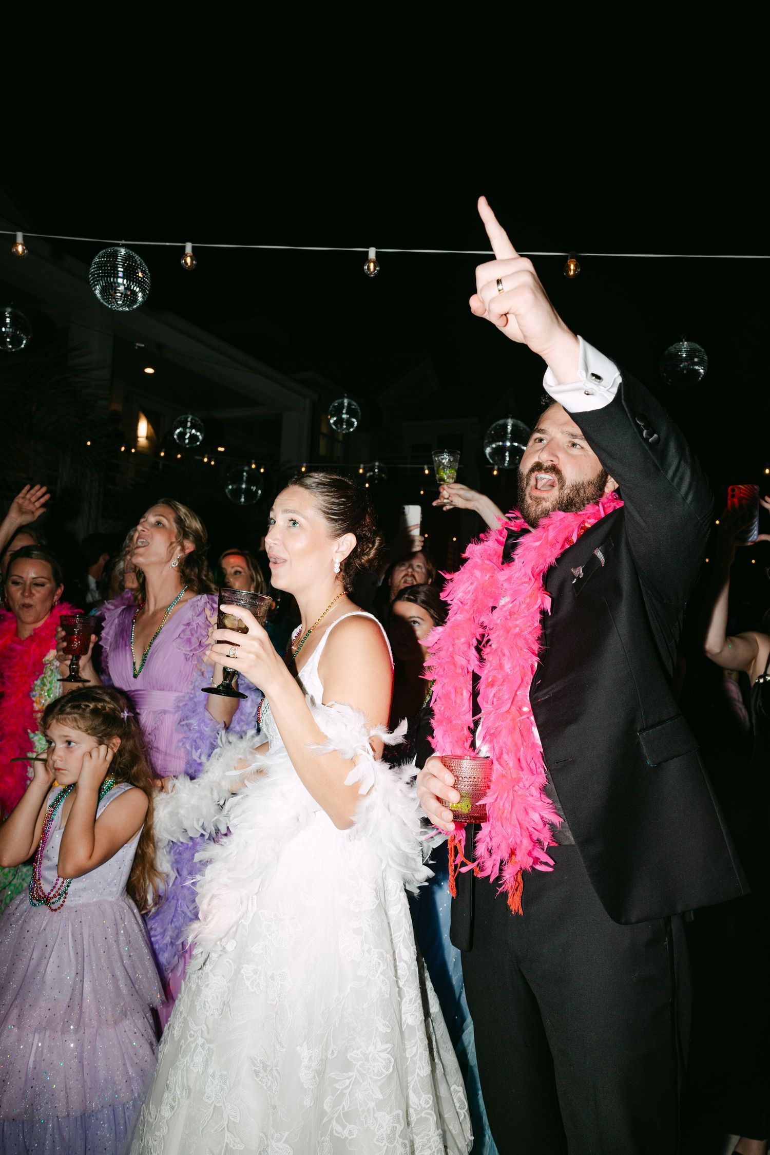 Bride and groom celebrate at wedding reception, groom wearing pink feather boa pointing up at disco ball lights.