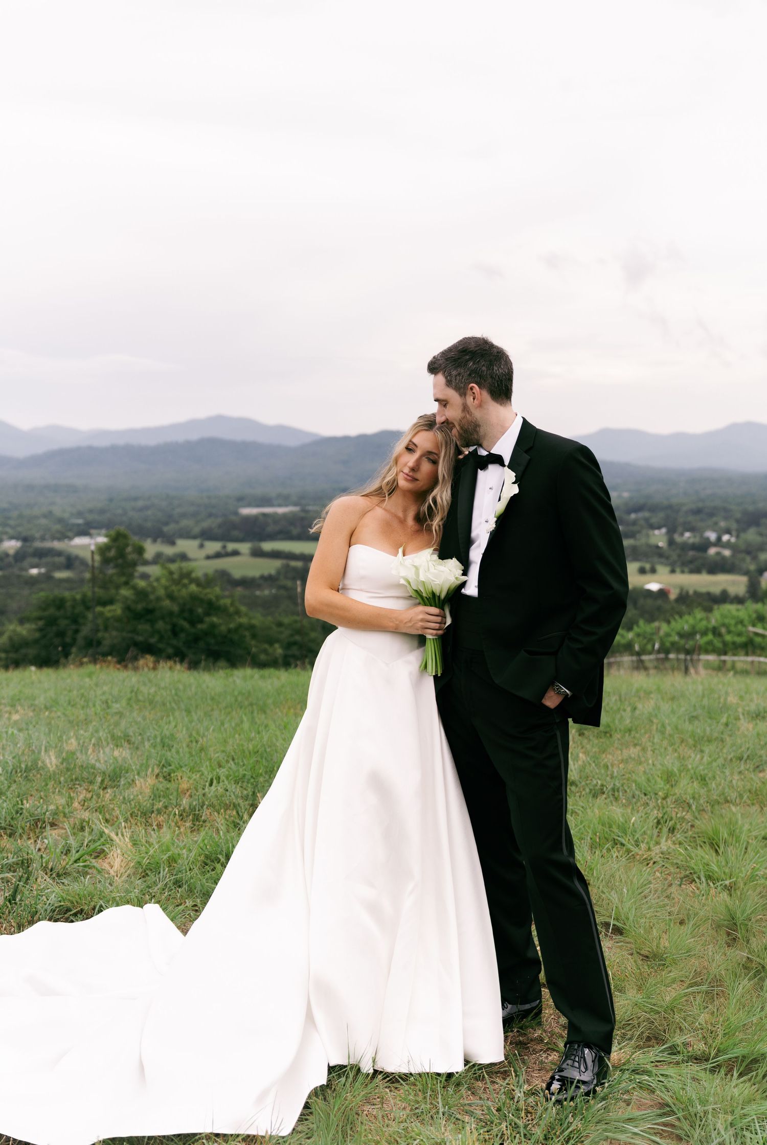 Bride in white strapless gown holds bouquet beside groom in black tuxedo, with rolling green hills and mountains behind.