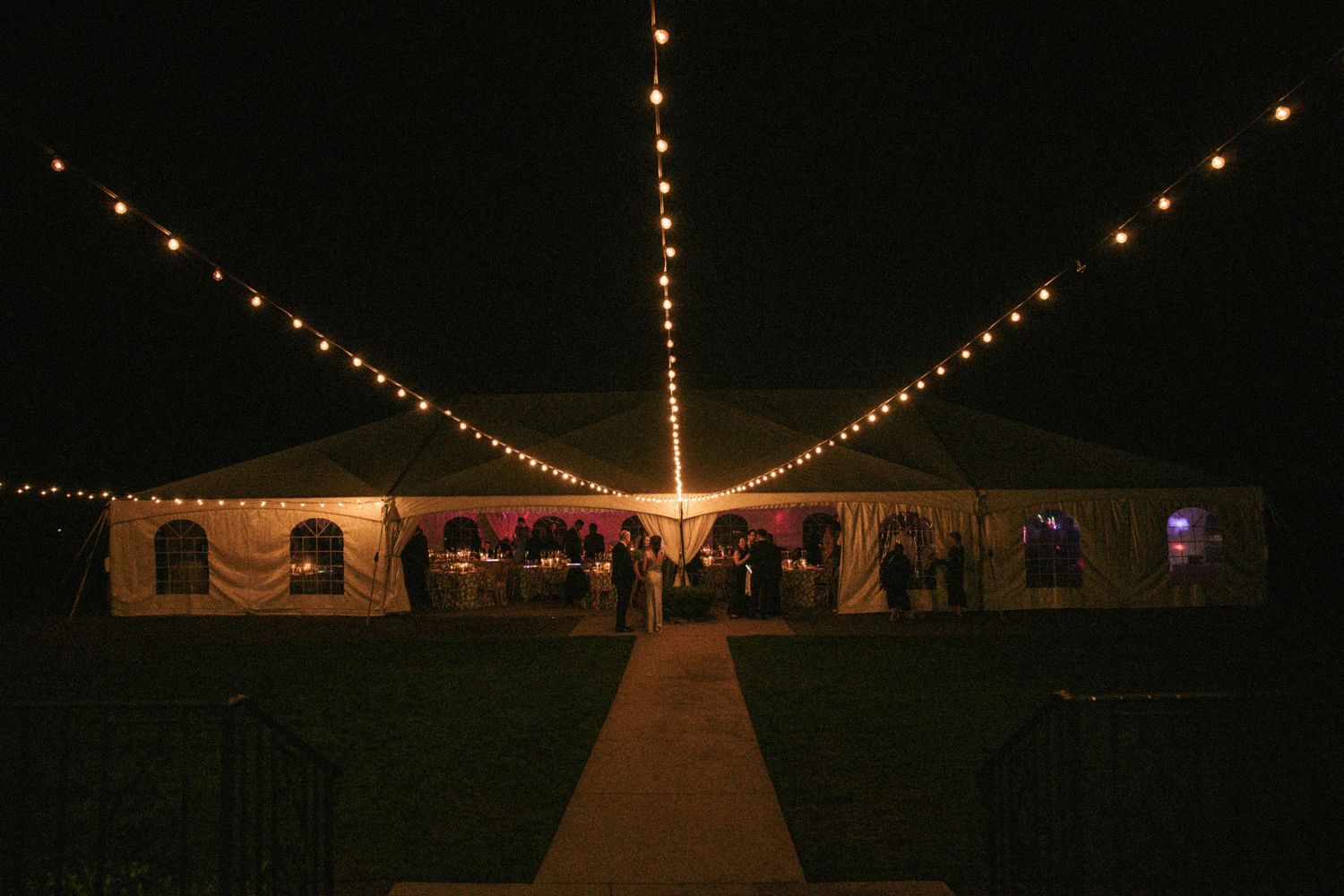 Large white event tent illuminated at night with string lights and guests gathered inside for an outdoor evening celebration.