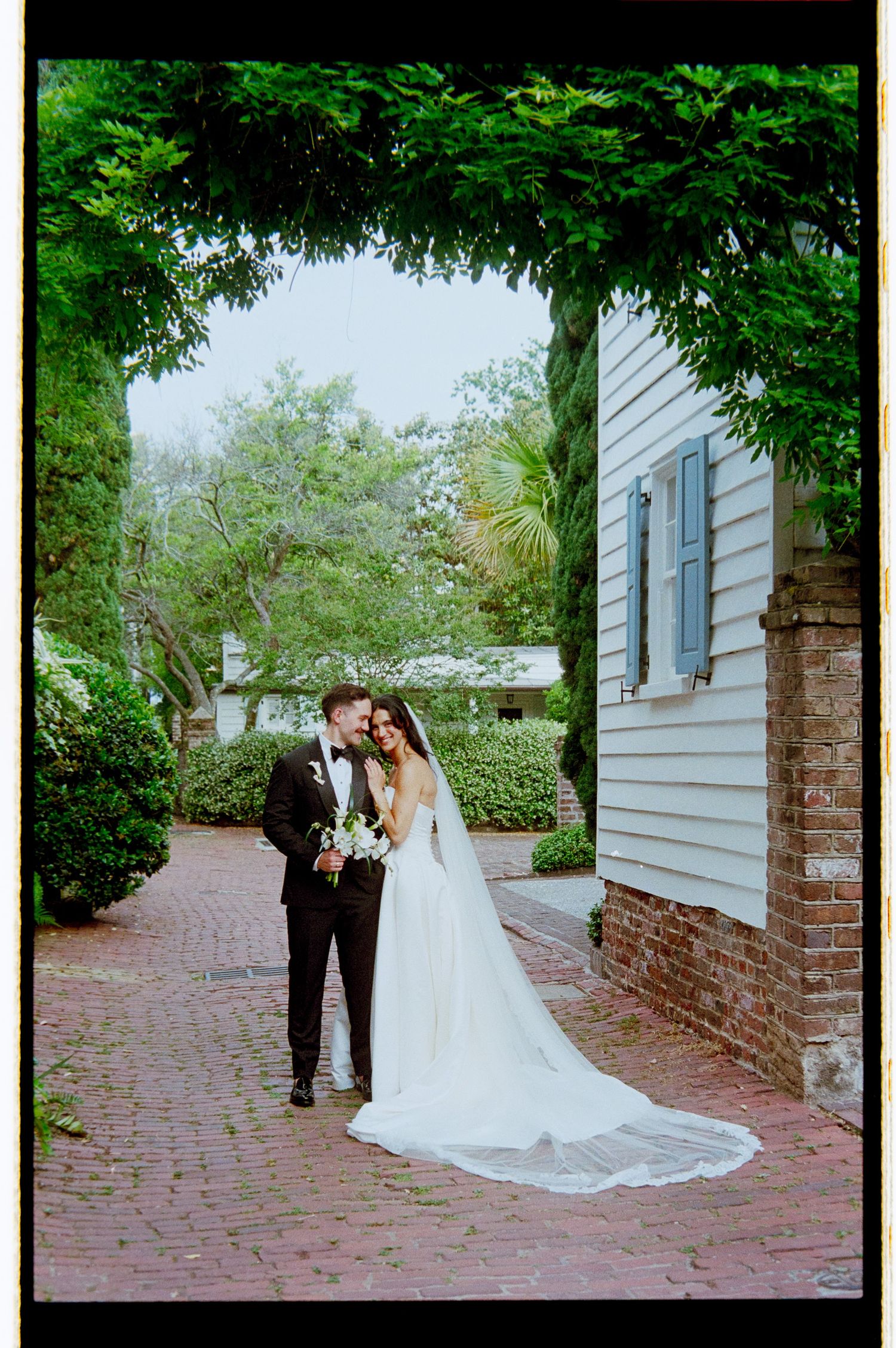 Bride and groom embrace on brick pathway beneath lush green archway in romantic film photography portrait.
