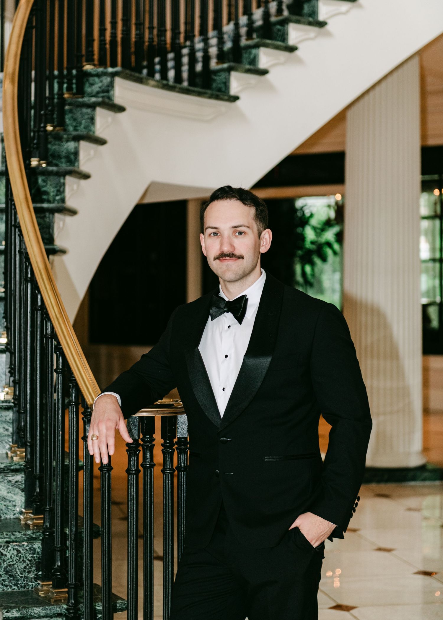 Dapper groom in black tuxedo with bow tie posing by a curved marble staircase with gold railing.