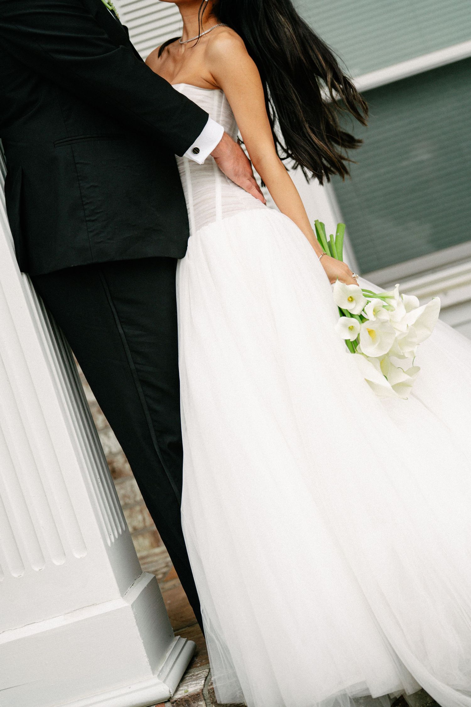 Bride in white strapless ball gown holding calla lily bouquet embraces groom in black tuxedo near white column.