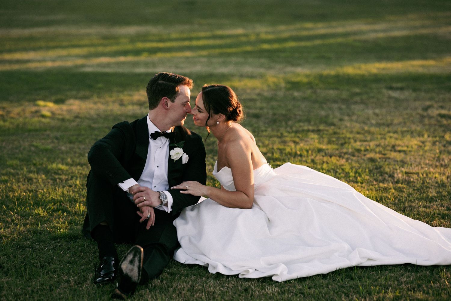 A bride and groom share a romantic kiss while sitting on a lush green lawn during their outdoor wedding celebration.