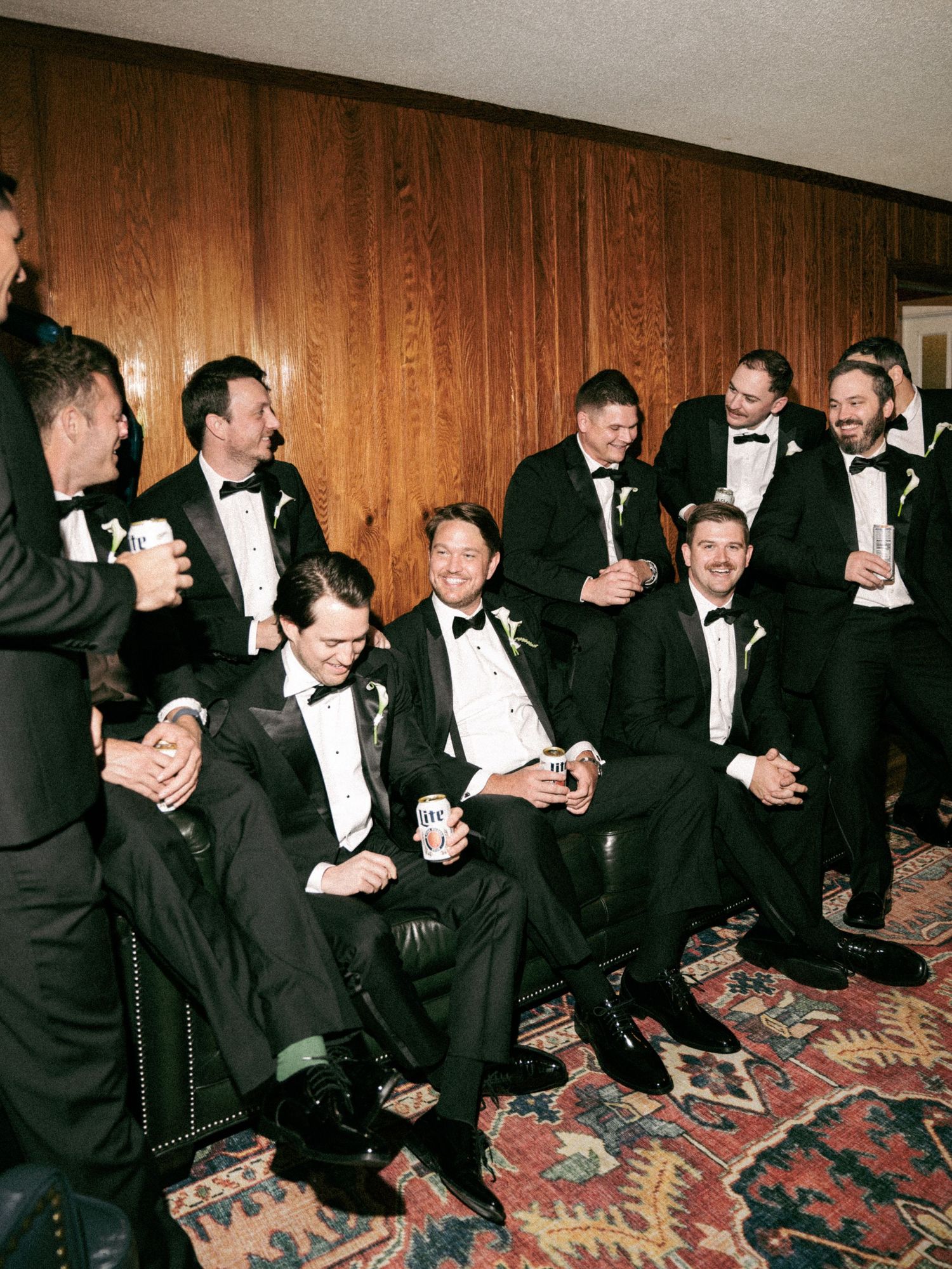 Groomsmen in black tuxedos laughing and celebrating together before a wedding ceremony.