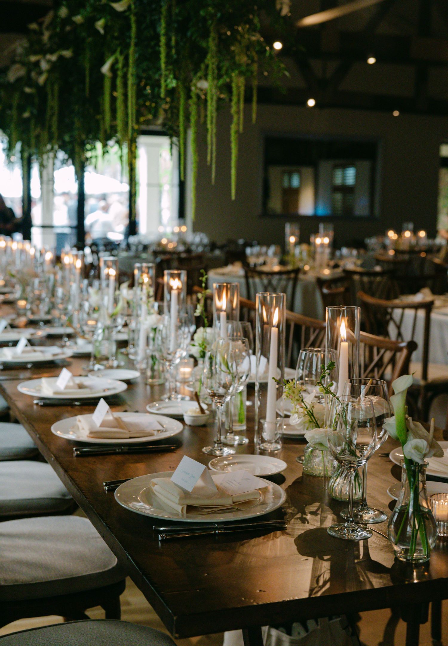 Long wooden dining tables set with crystal glasses and white plates under hanging greenery decor.