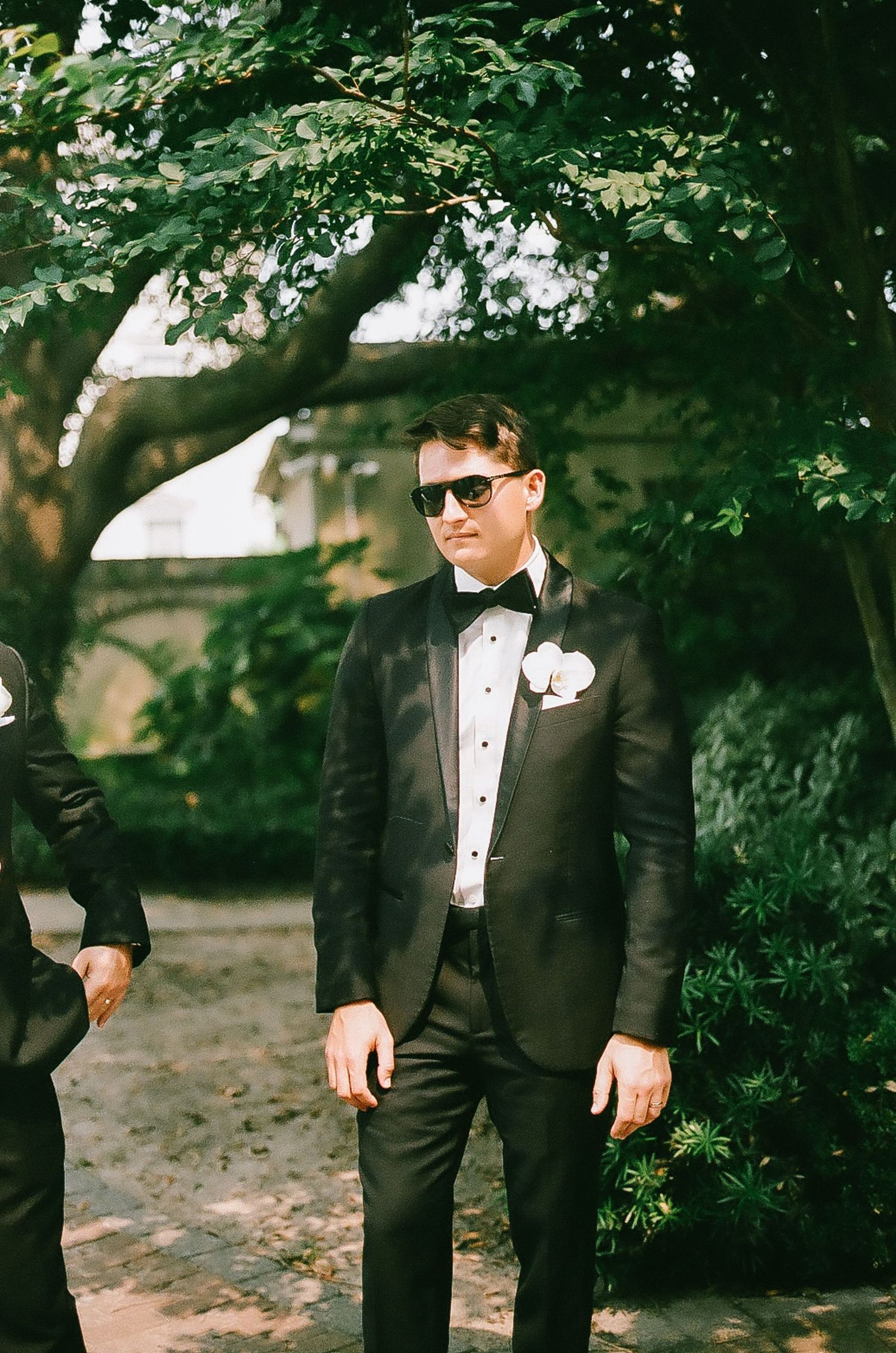 Groomsmen in black tuxedos and sunglasses pose casually outdoors under tree canopy.
