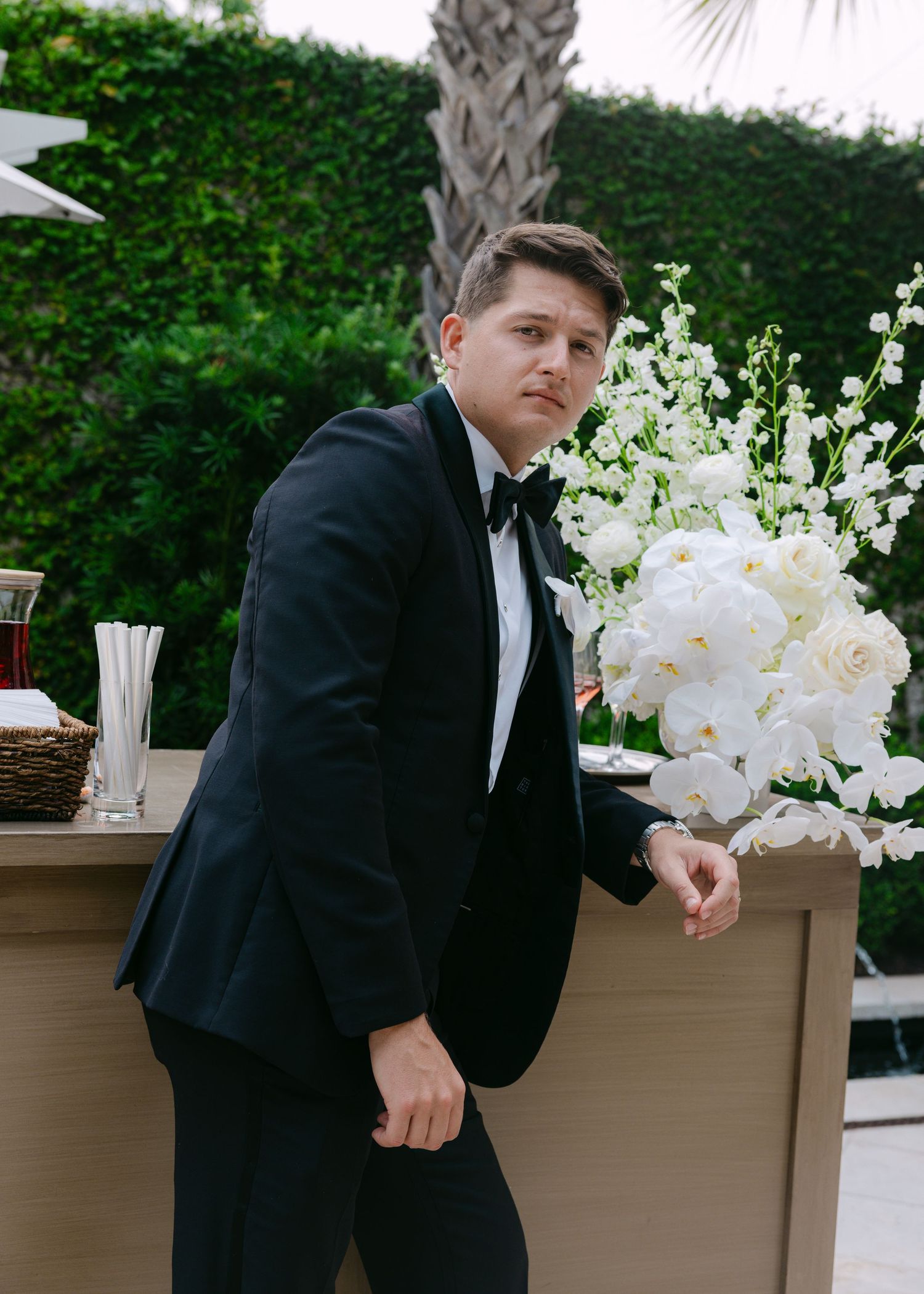 Groom in classic black tuxedo poses playfully in front of garden planters with white flowers.