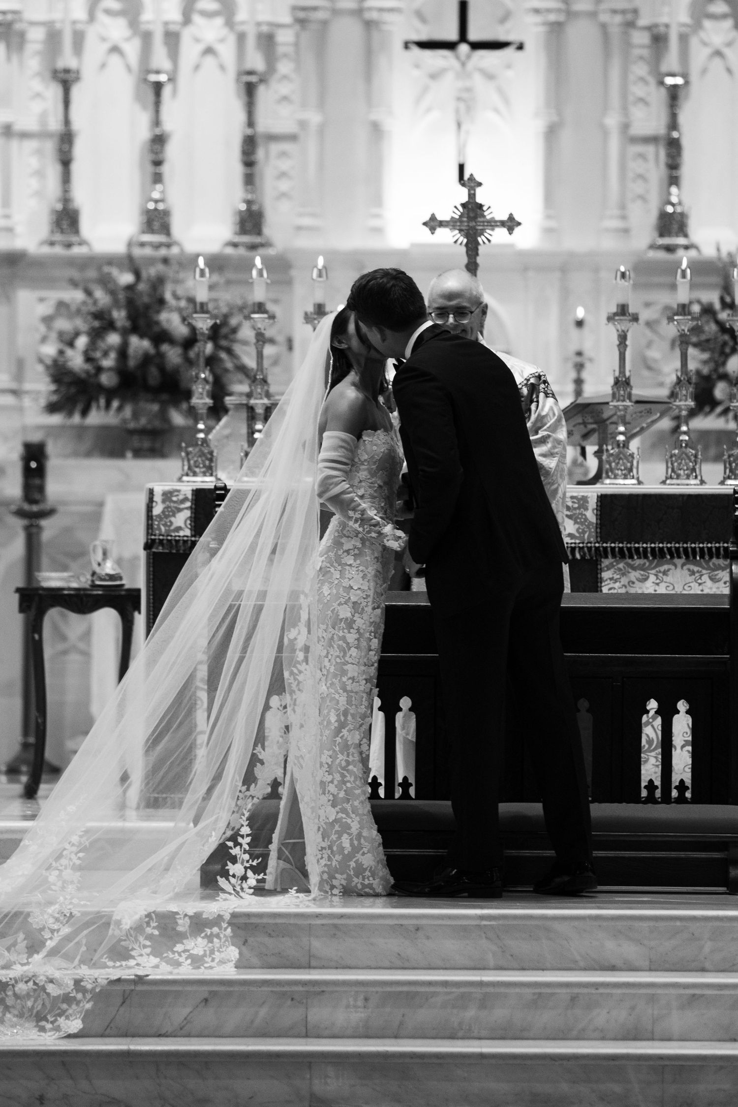 Black and white photo of a couple at the altar with a long lace veil trailing on cathedral steps.
