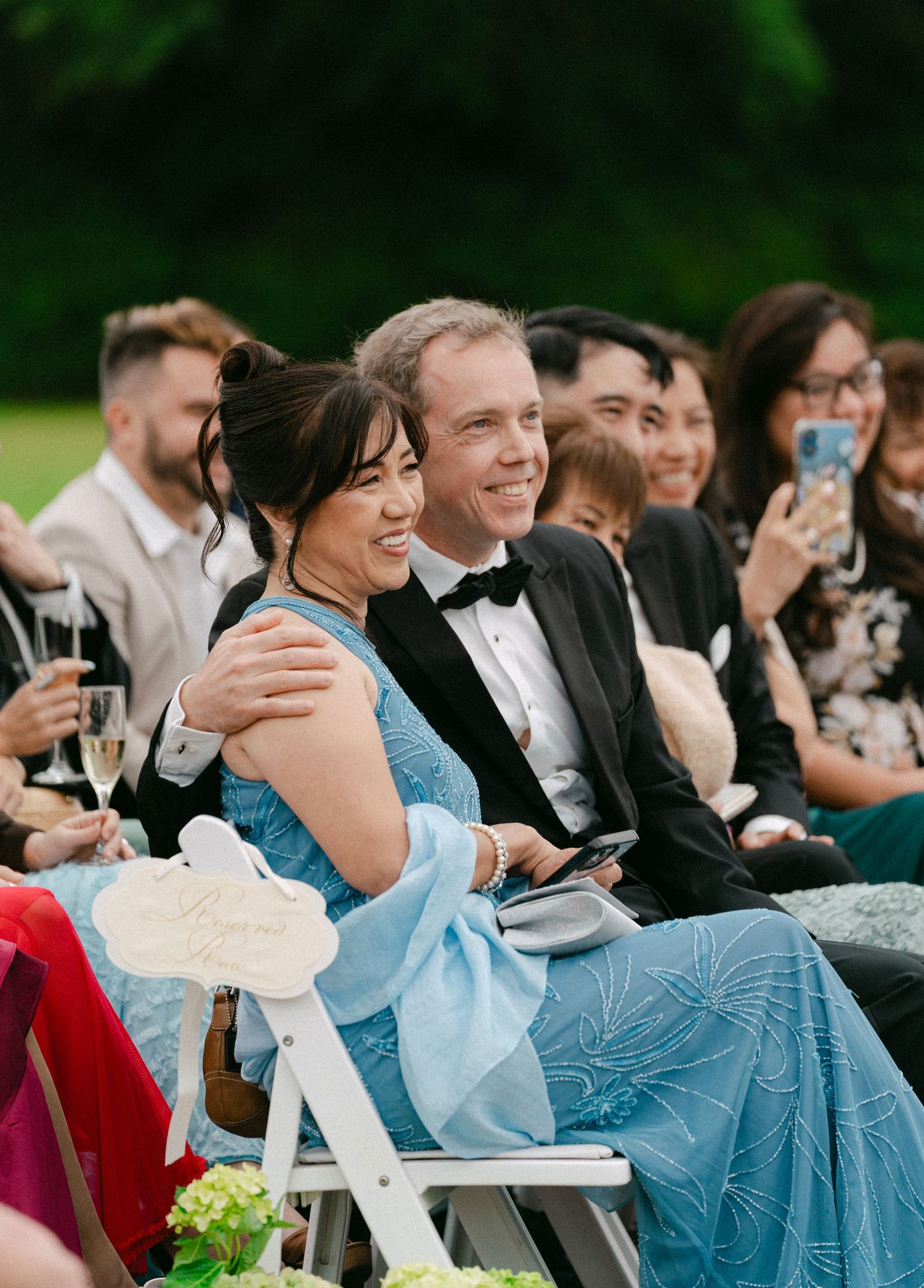 Smiling wedding guests seated outdoors, woman in blue dress embraced by man in tuxedo during ceremony.
