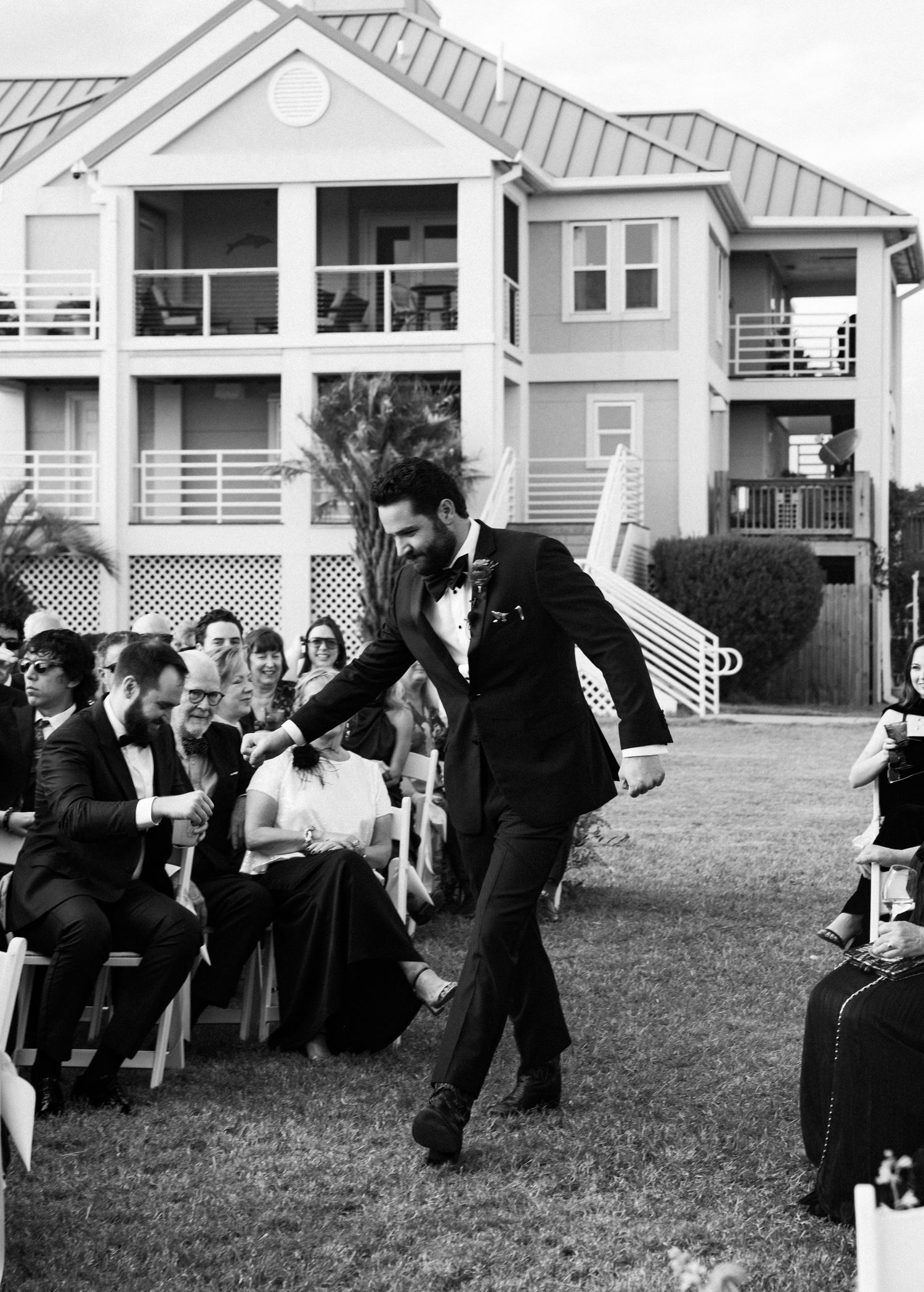Groom in black tuxedo walks down outdoor wedding ceremony aisle in black and white photo.