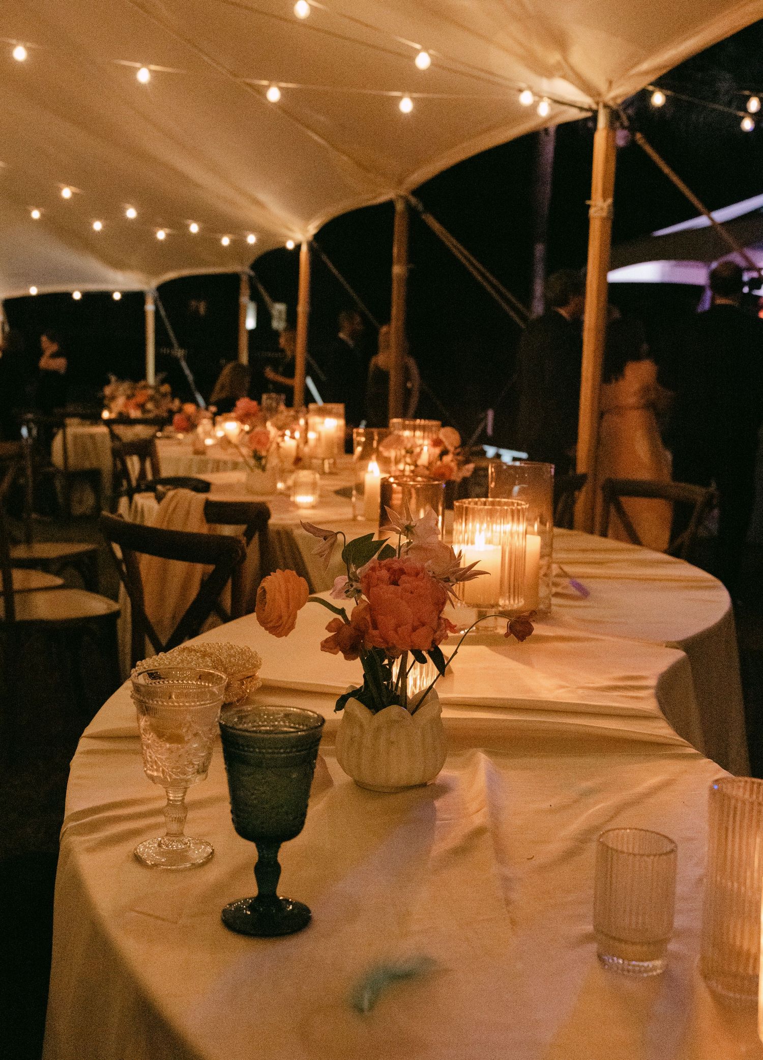 Romantic wedding reception under a tent with string lights, candlelit tables, floral centerpieces, and elegant glassware at night.