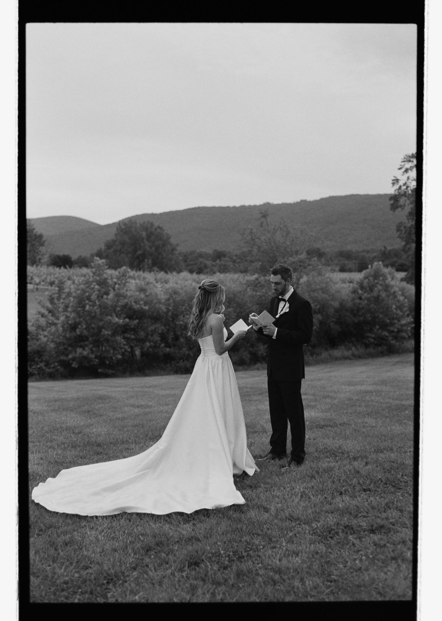 Black and white film photo of bride and groom exchanging vows on a lawn with mountain backdrop.