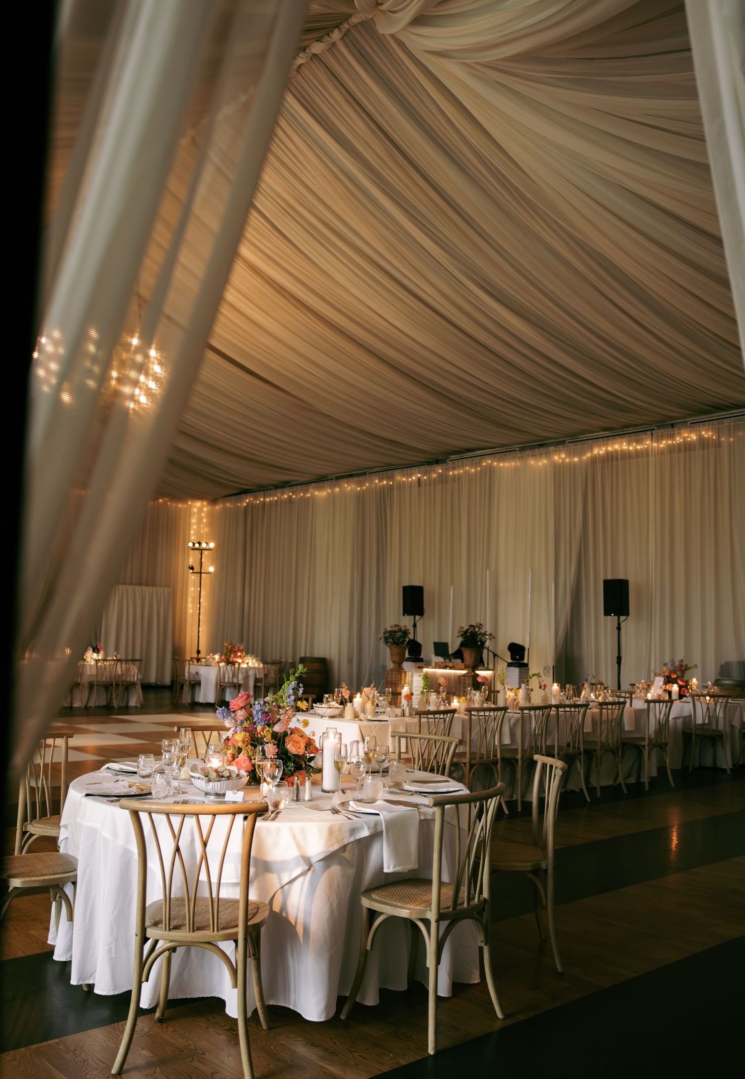 Romantic tented wedding reception with draped ceiling, string lights, round tables, and gold chairs.