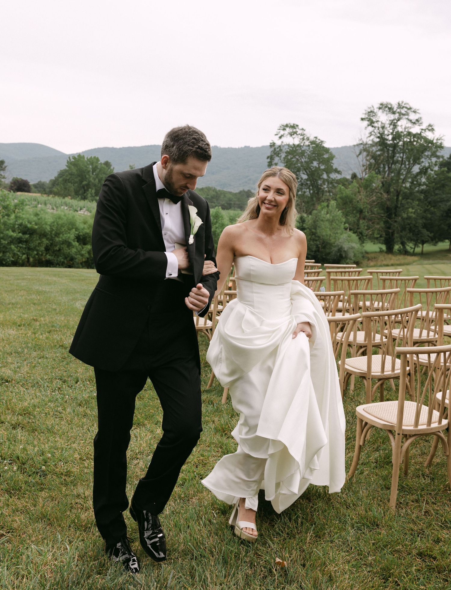 Bride in strapless white gown and groom in black tuxedo walk joyfully past wooden chairs at outdoor wedding ceremony.