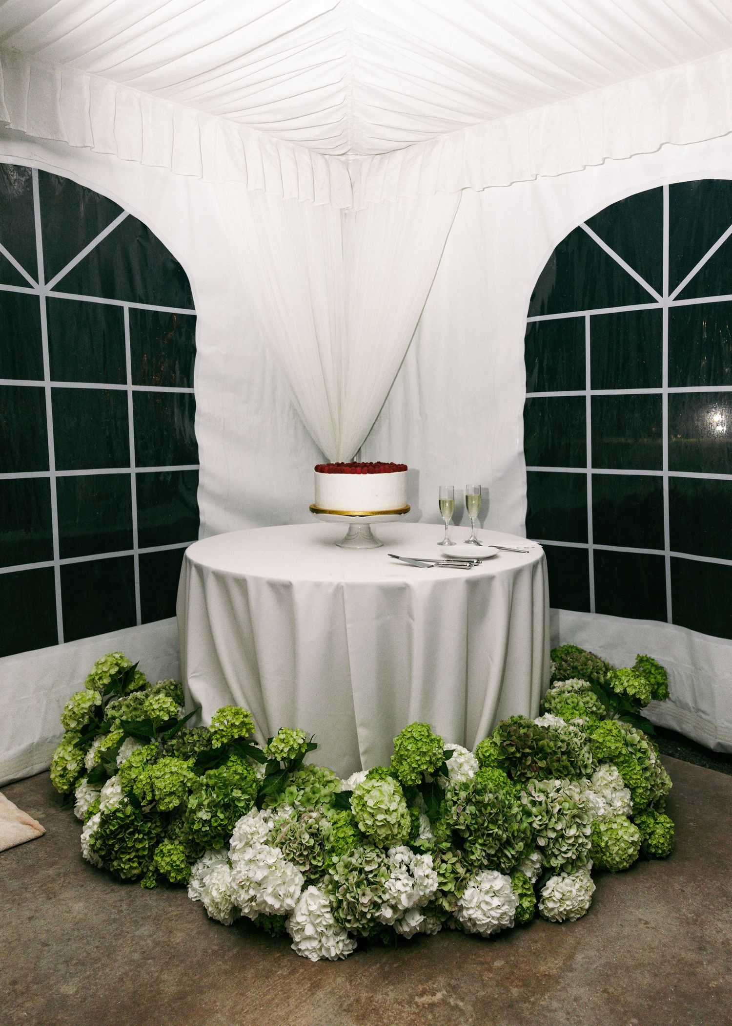 Wedding cake on a round table surrounded by lush green and white hydrangea floral arrangements inside a white tent.