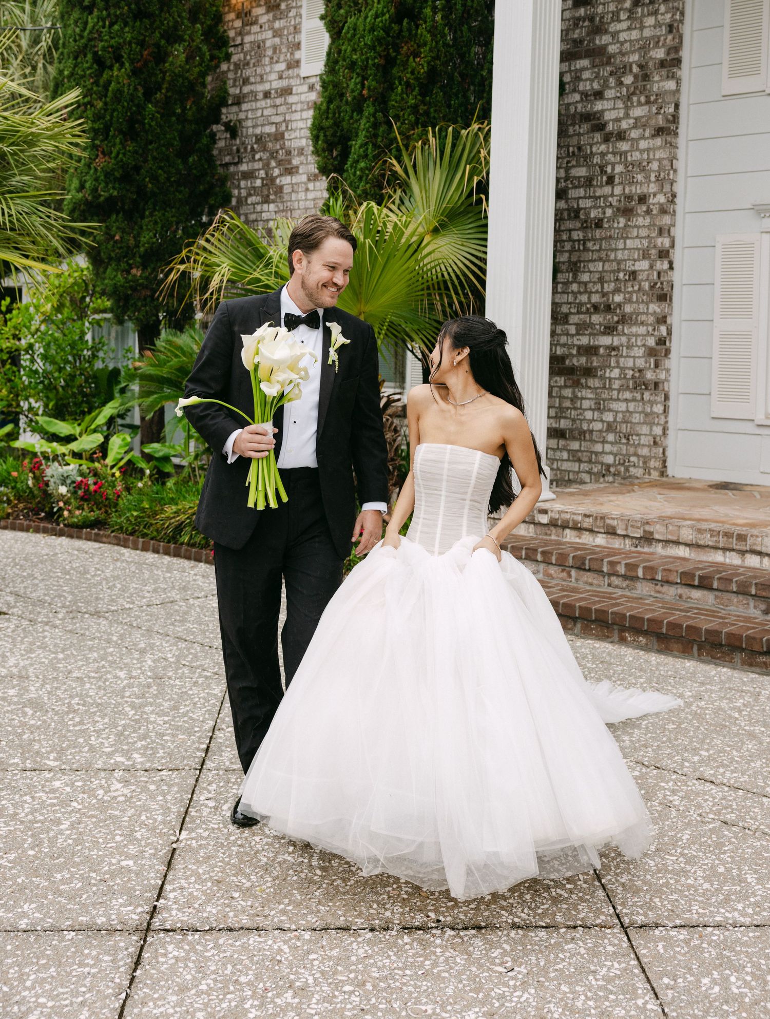 A bride in a strapless tulle ball gown and groom in black tuxedo walk together outside a brick building.
