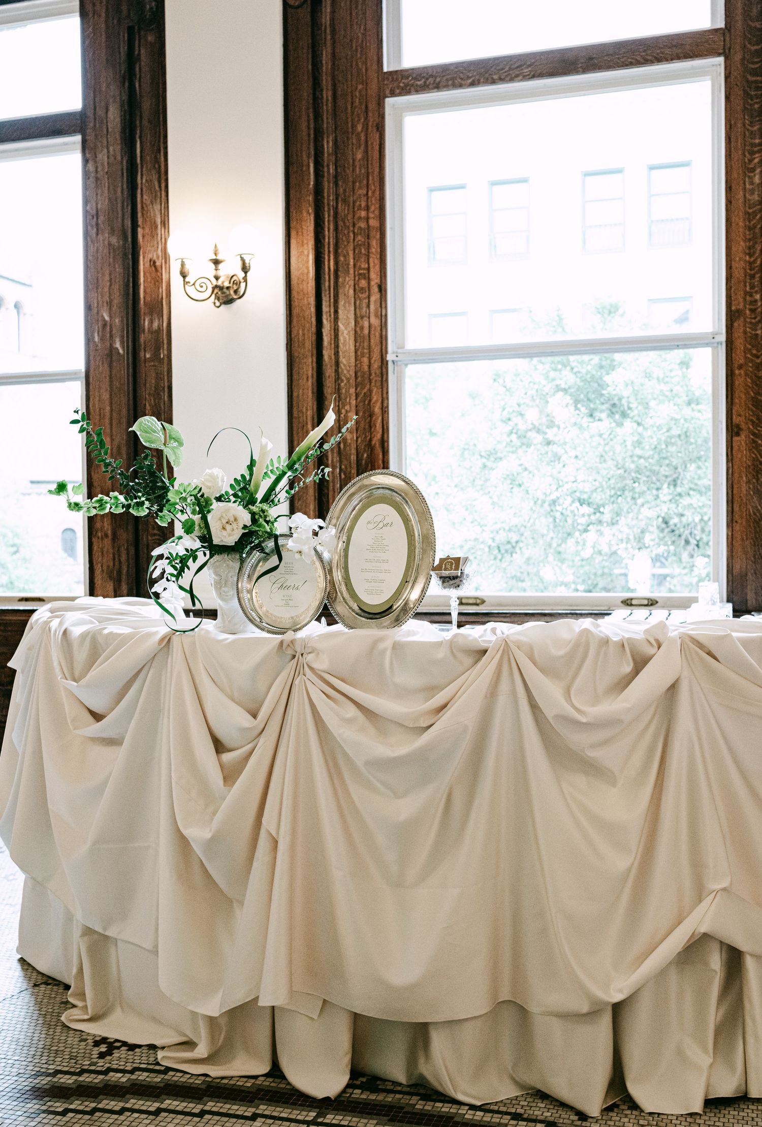 Elegant wedding reception table with draped ivory fabric, floral arrangements, and framed sign by tall windows.