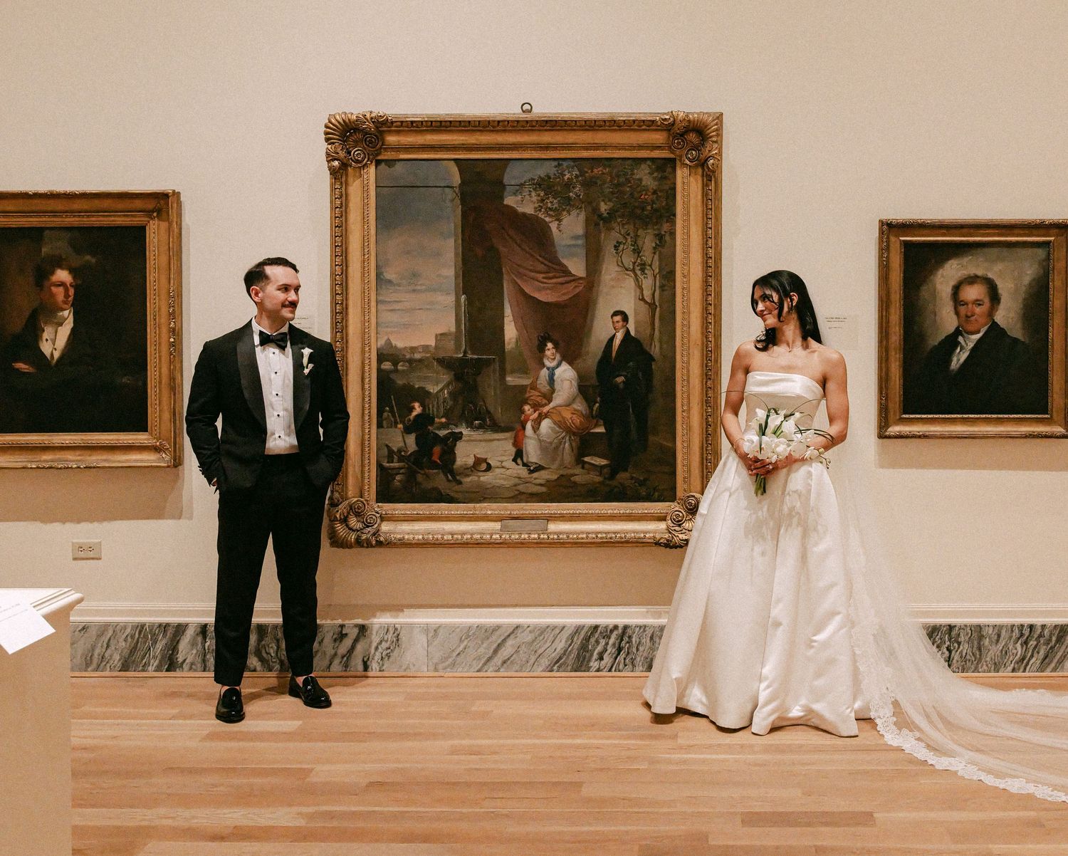 Bride and groom posing apart in an art museum gallery surrounded by classic portrait paintings on the walls.