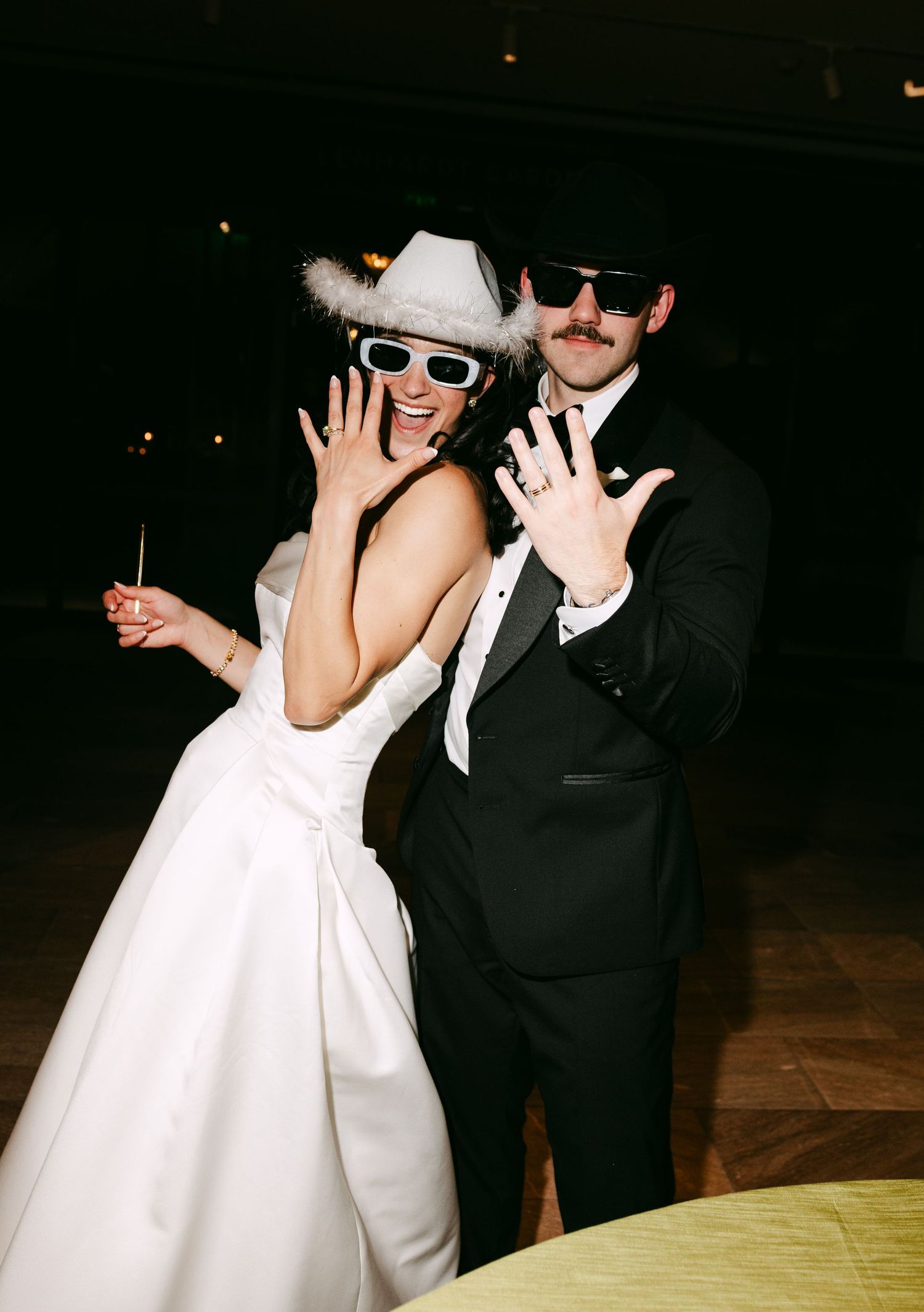 Newlyweds in sunglasses and cowboy hat showing off wedding rings at fun reception celebration.