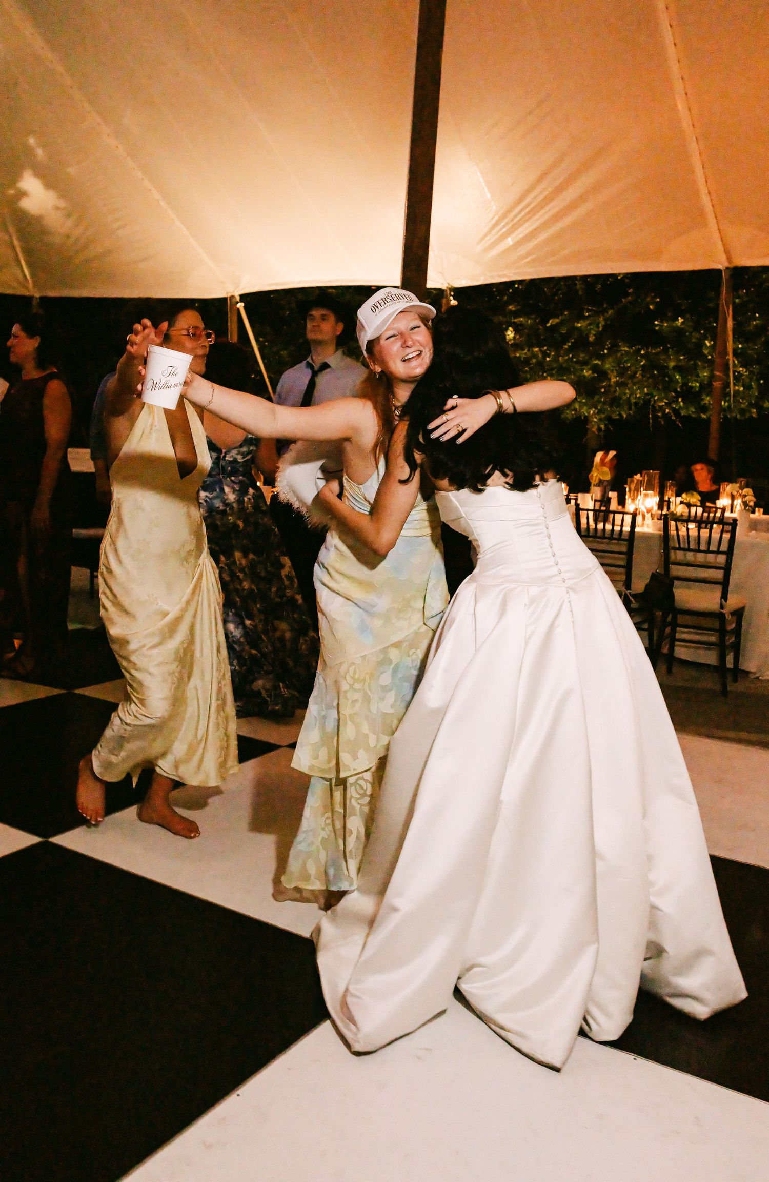 Bride in white strapless ball gown dances joyfully with guests on black and white checkered dance floor.