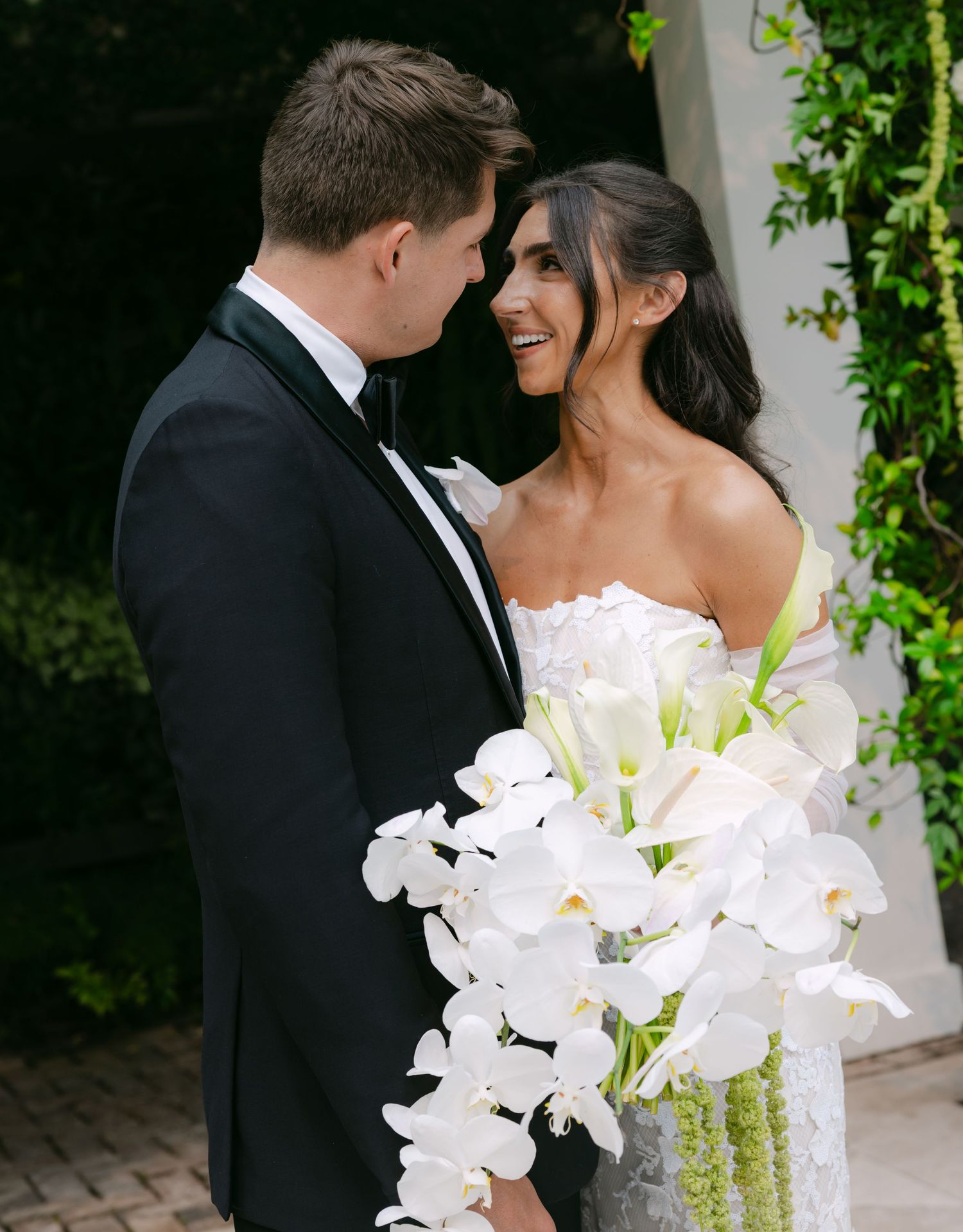 Couple in wedding attire stands close together with a stunning white orchid bouquet against a green garden backdrop.