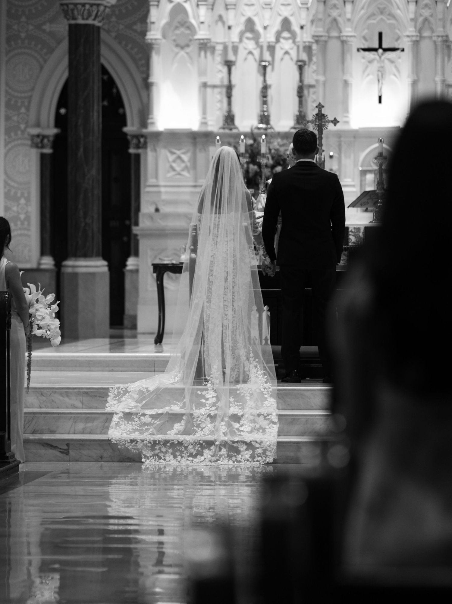Black and white photo of a wedding ceremony in a cathedral with a long lace veil trailing behind.