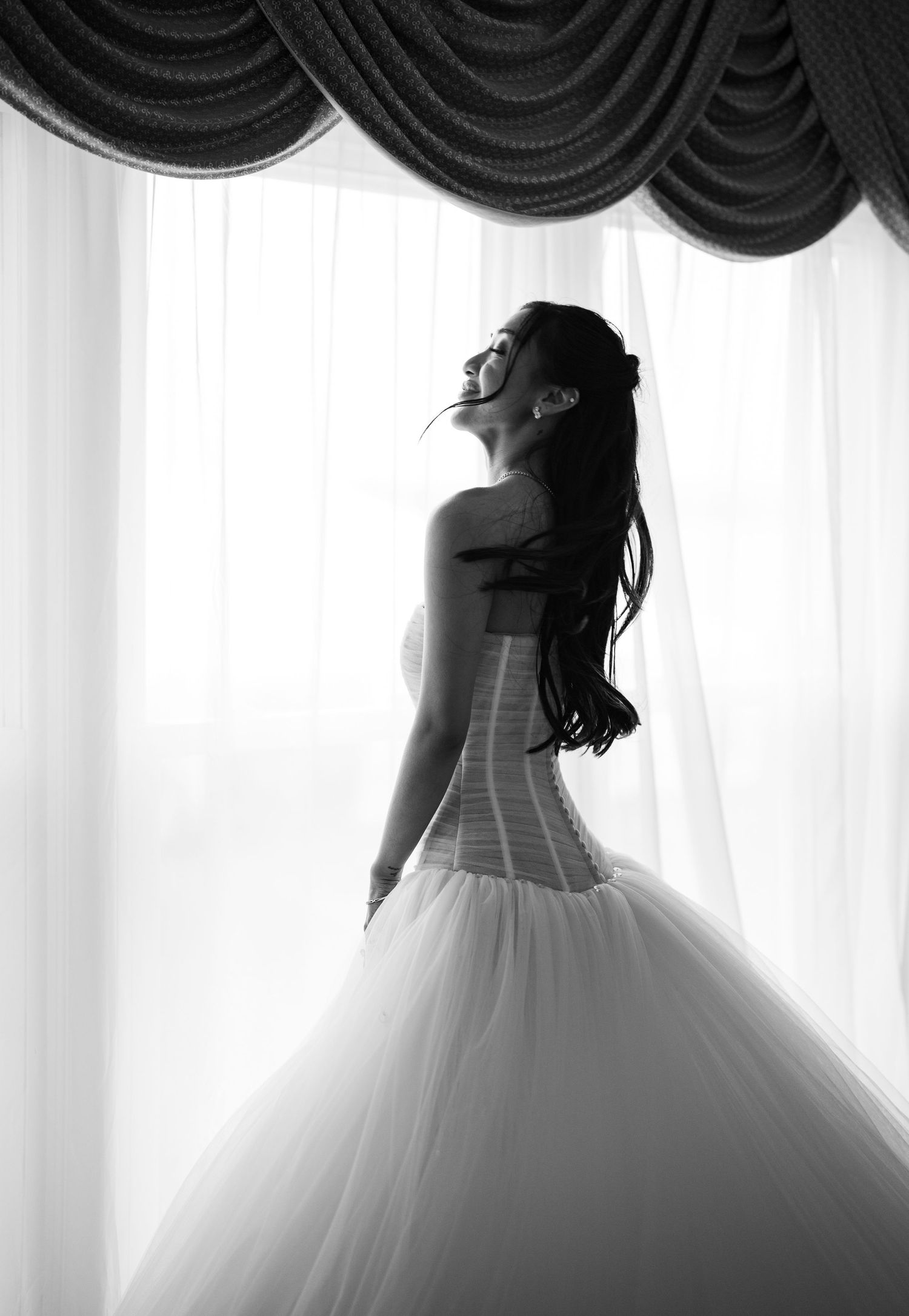 Black and white silhouette of a bride in a ballgown standing by sheer curtained windows.