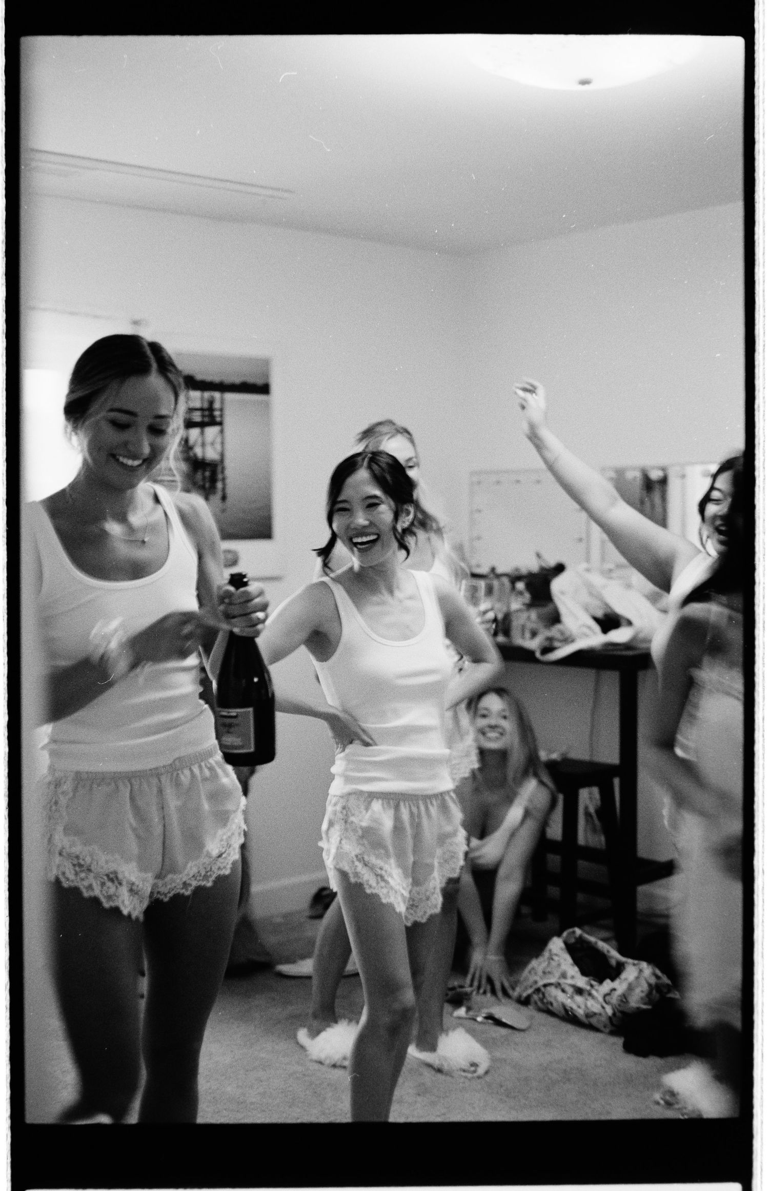 Women in white tank tops and lacy shorts celebrating with champagne in a black and white photo.