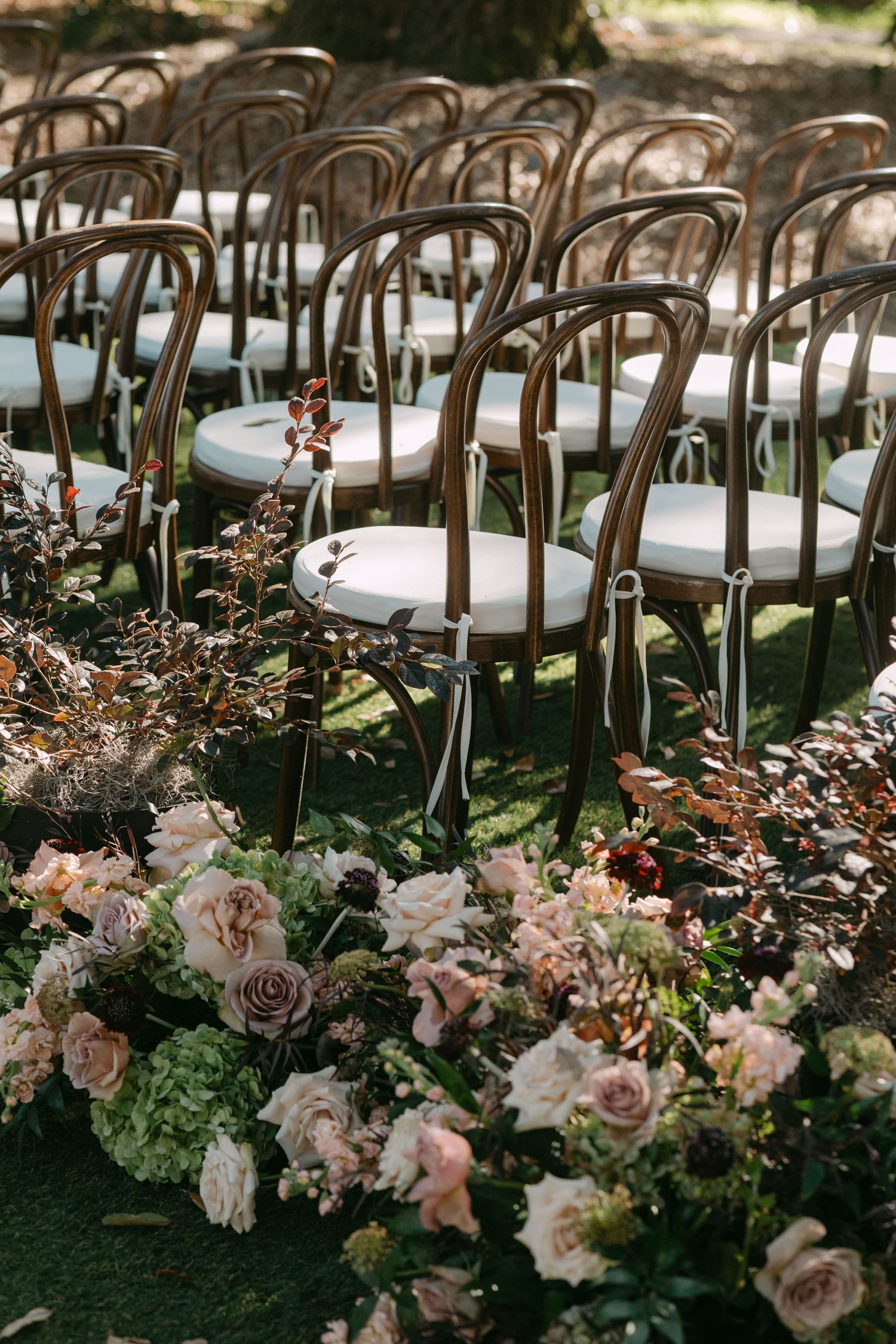 Elegant wedding ceremony setup with antique bronze chairs lined up and decorated with blush roses and greenery along the aisle.