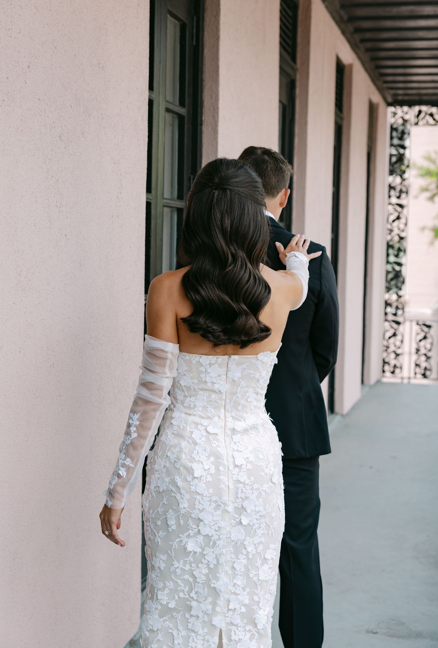 Back view of a bride in a form-fitting lace dress following her groom along a covered walkway.