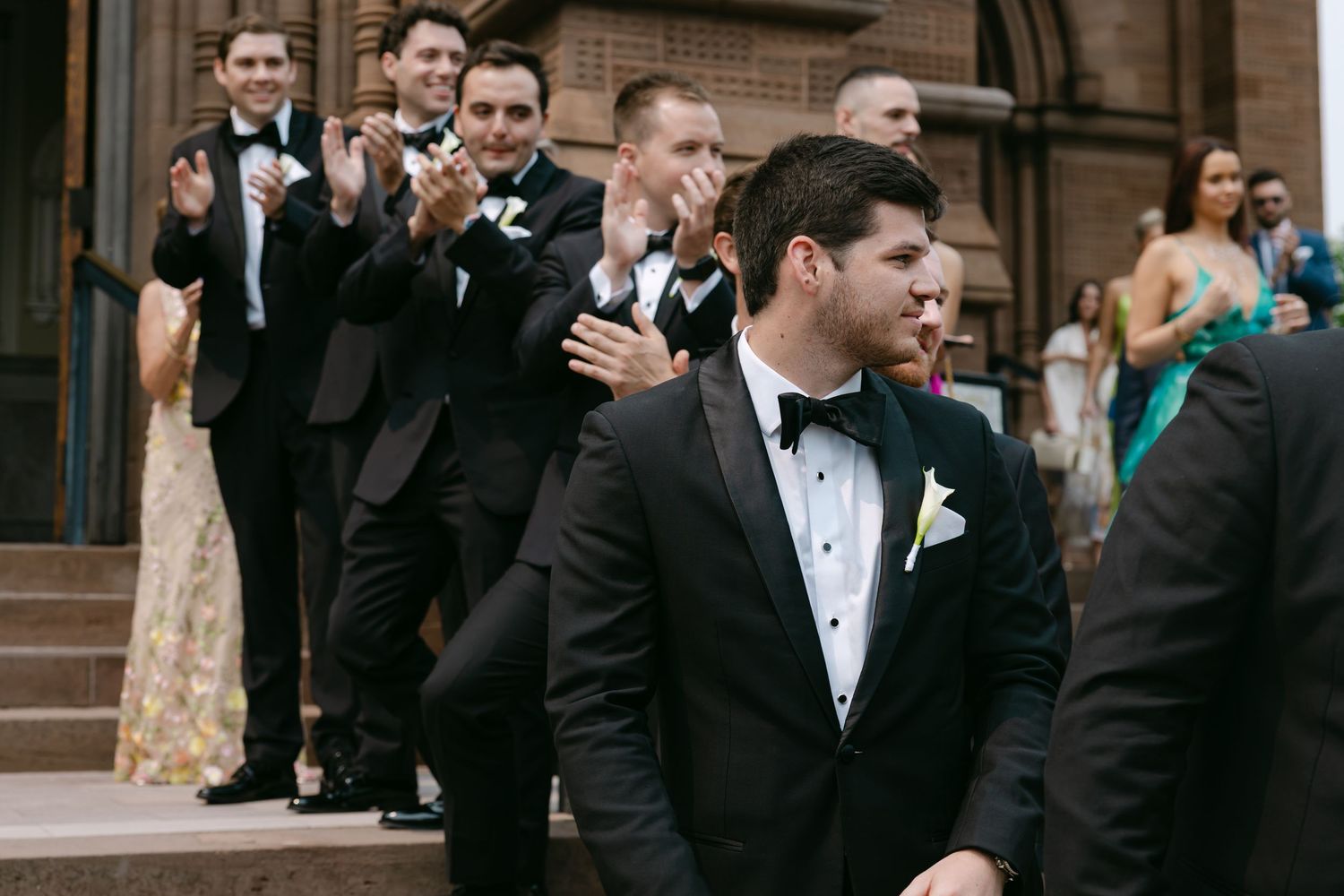 Groomsmen in black tuxedos gather outside church entrance.