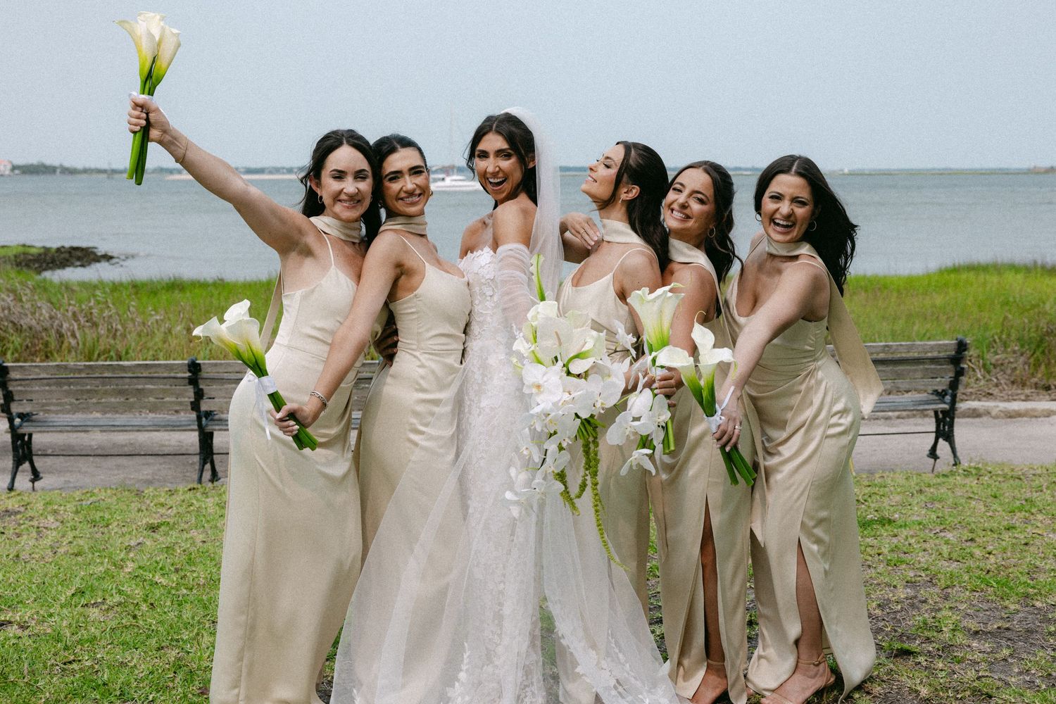 Wedding party in champagne dresses poses playfully on a waterfront lawn.