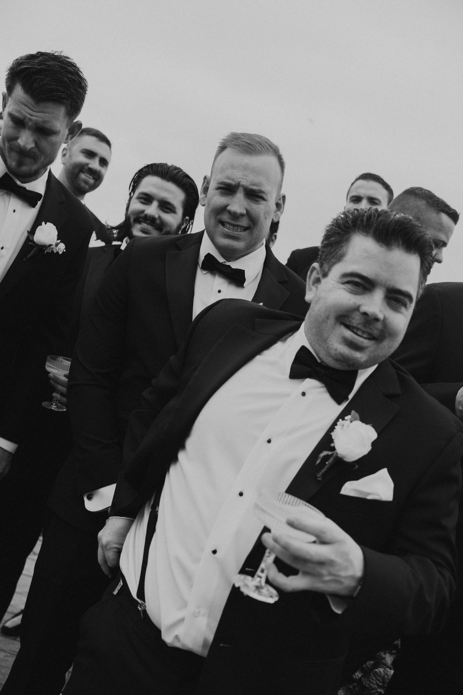 Group of groomsmen in black tuxedos and bow ties laughing together in black and white photo.